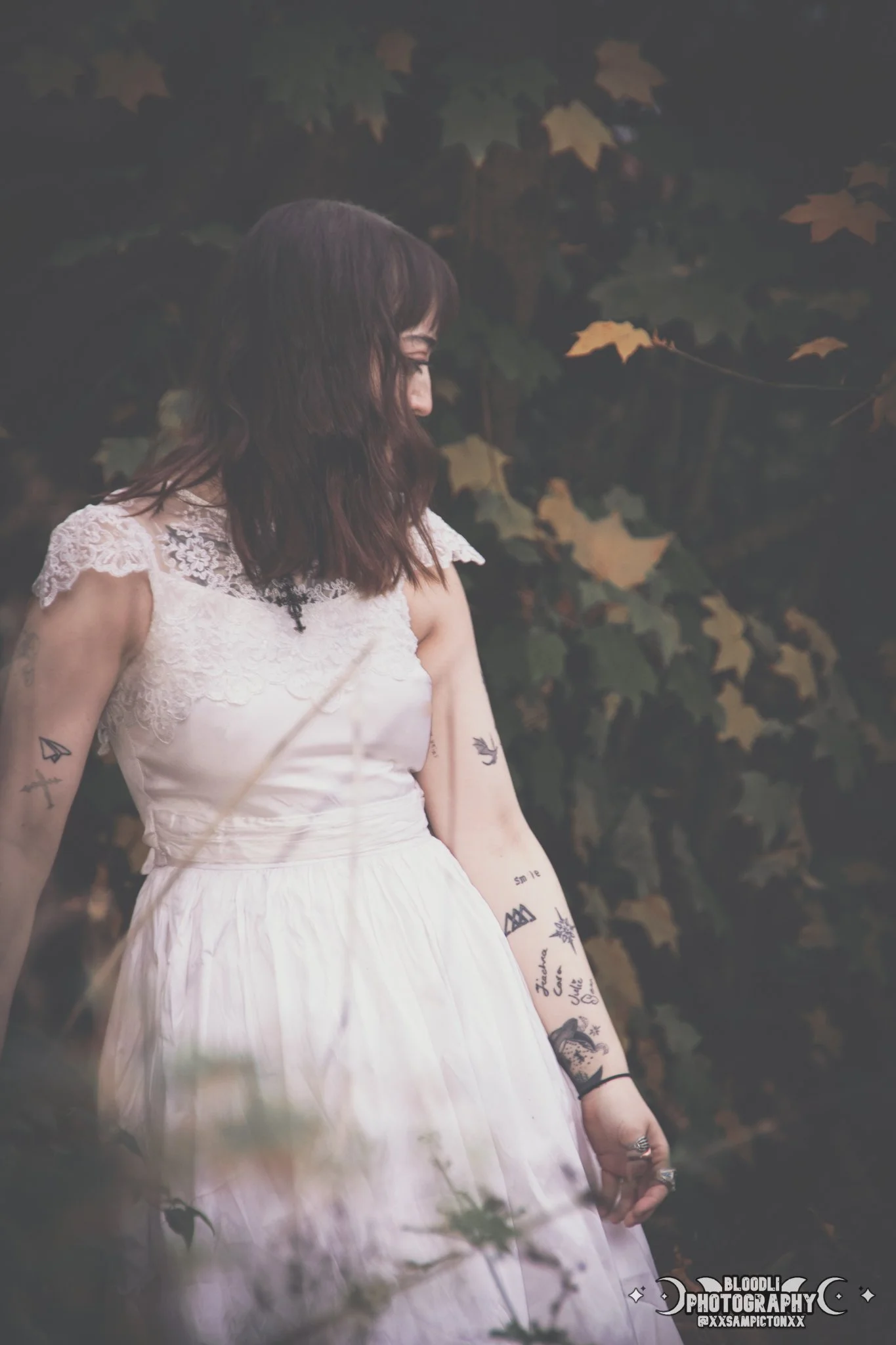 A woman with dark brown, shoulder-length hair is wearing a white lace dress. She is standing outdoors amid greenery and autumn leaves, looking down with a contemplative expression.