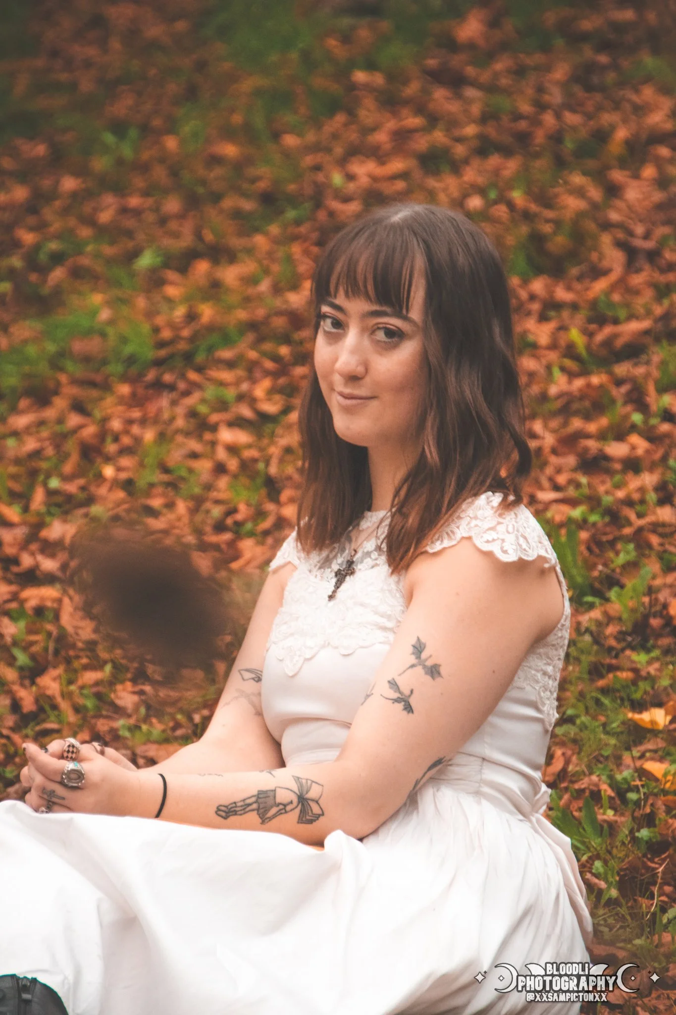 A young woman with dark brown hair and tattoos on her arms sitting outdoors on fallen leaves, wearing a white dress with lace details and a black necklace, looking at the camera with a slight smile.