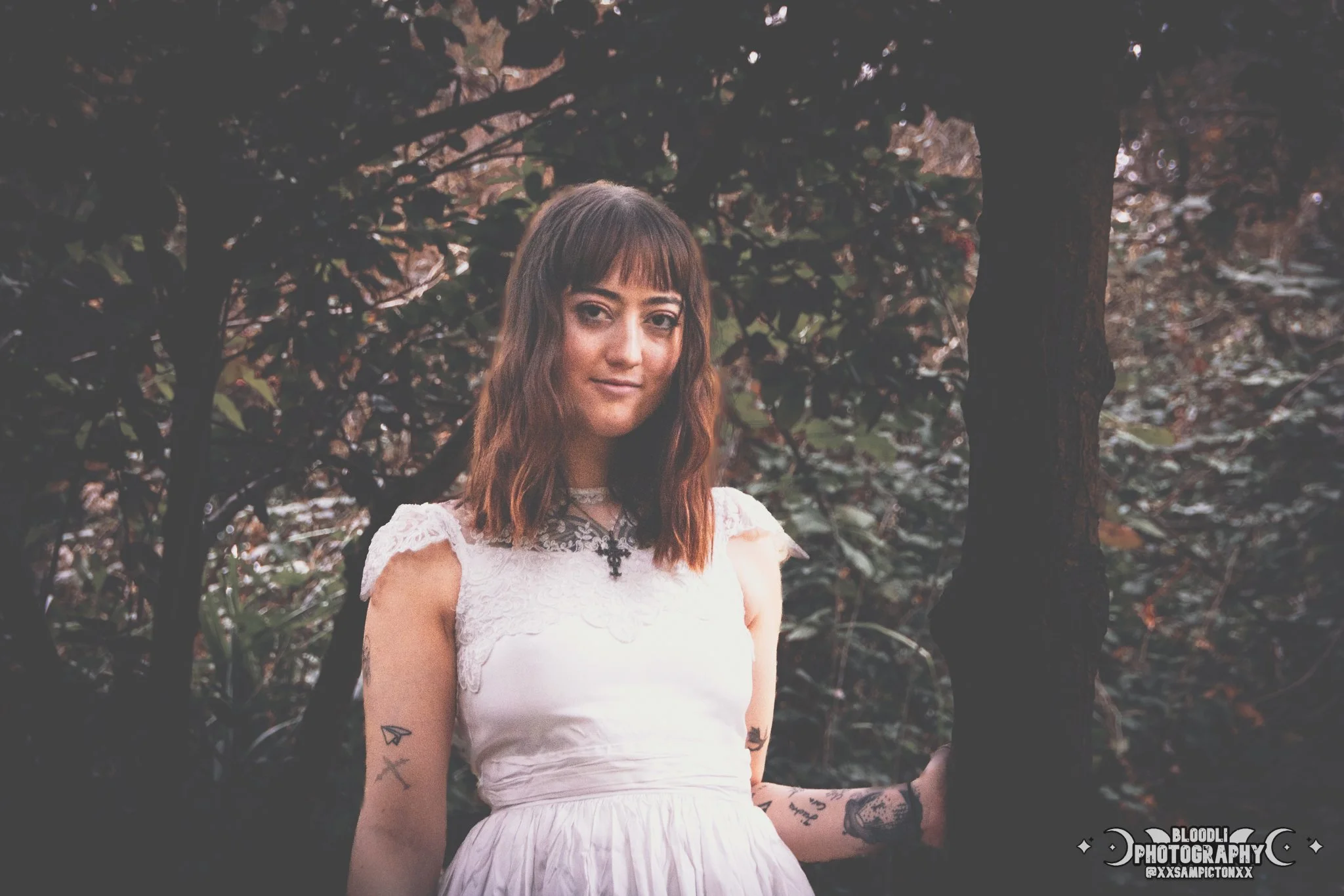 A young woman with shoulder-length brown hair and tattoos on her arms, wearing a white dress with lace details, is standing outdoors among trees with dark leaves. She is looking at the camera with a slight smile. The photo has a dim, moody lighting.