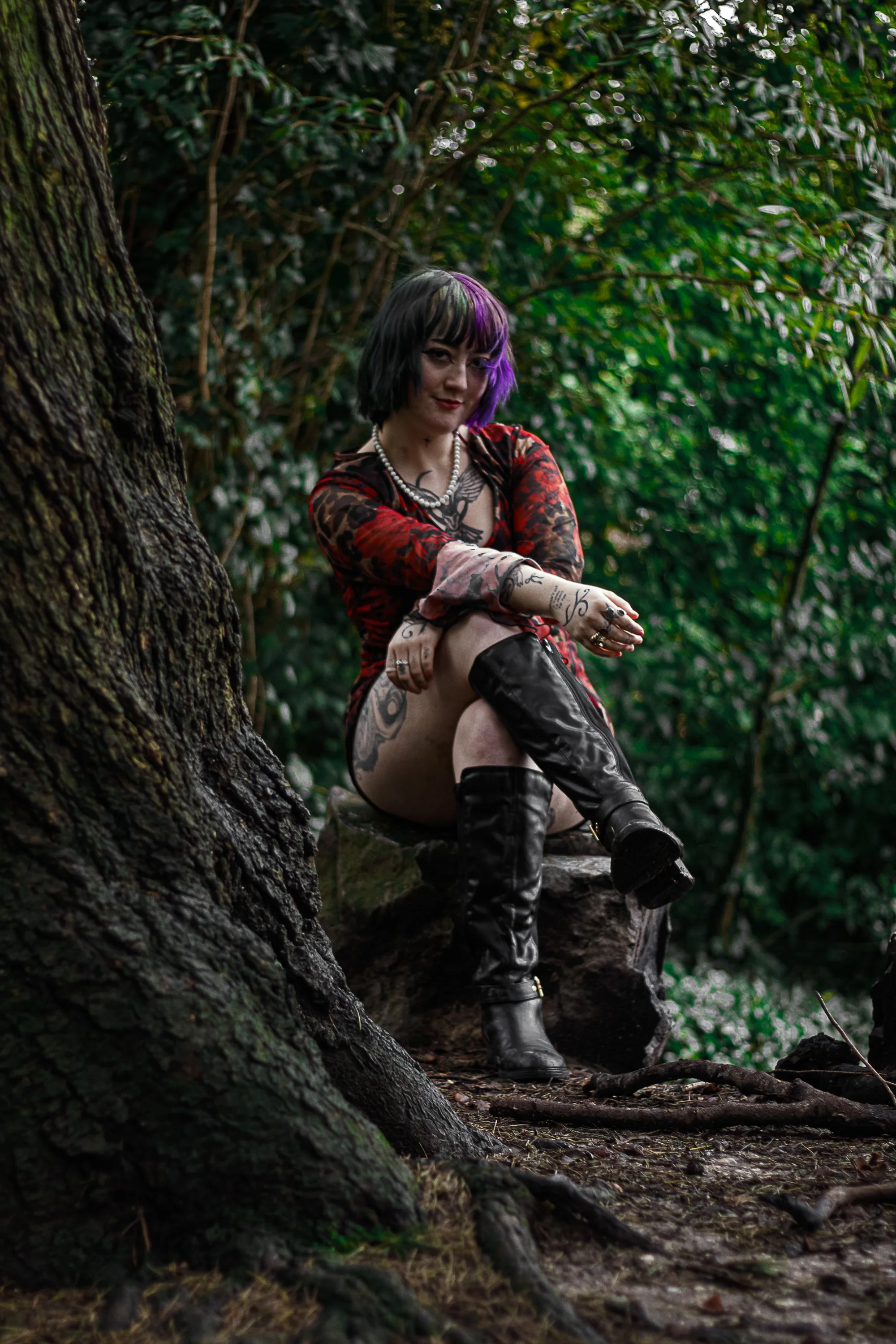 A woman with tattoos and purple and black hair sitting on a rock in a forest, wearing a red and black dress and knee-high black boots, looking at the camera with a confident expression.