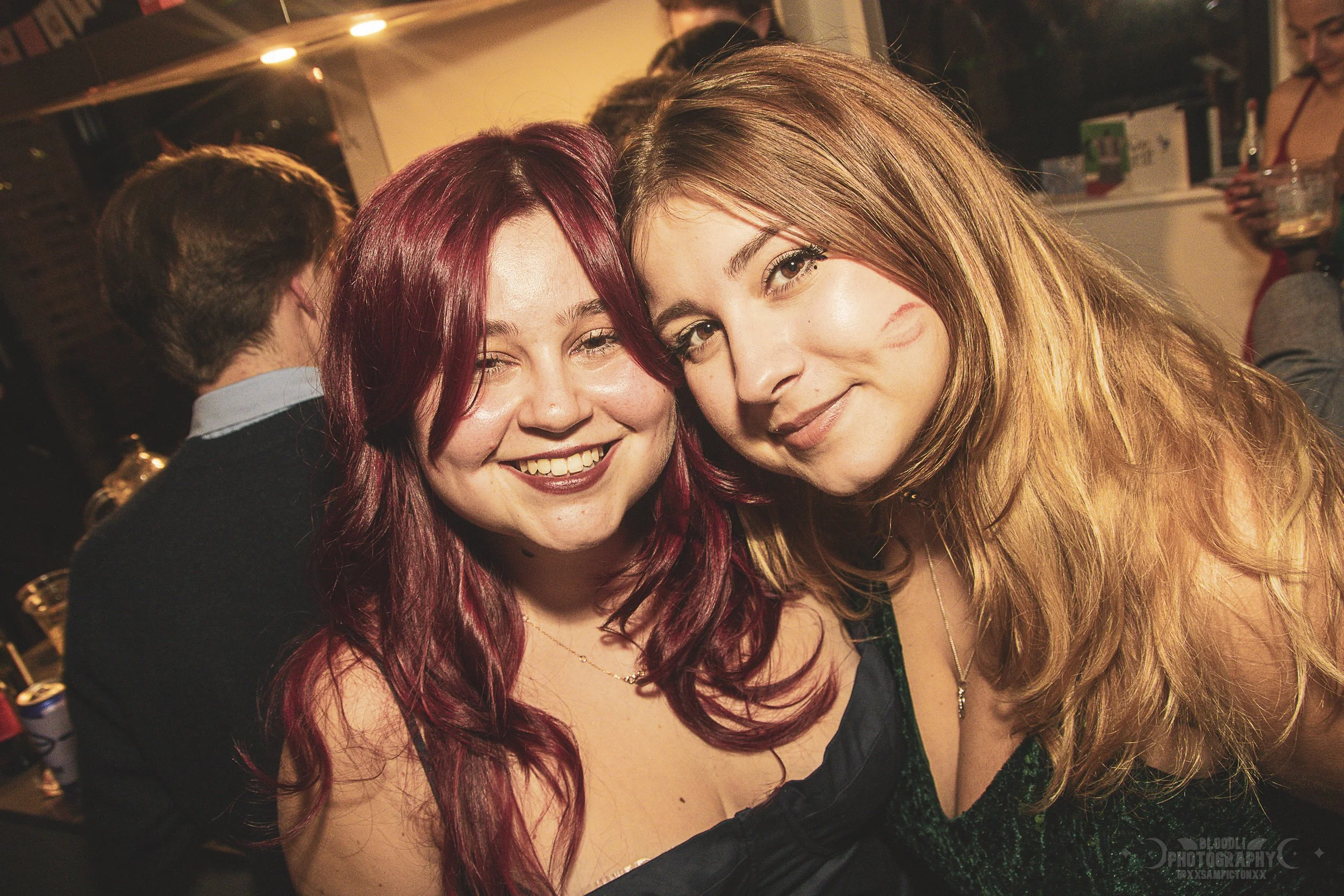 Two women smiling and hugging at a party or social gathering, with other people in the background.