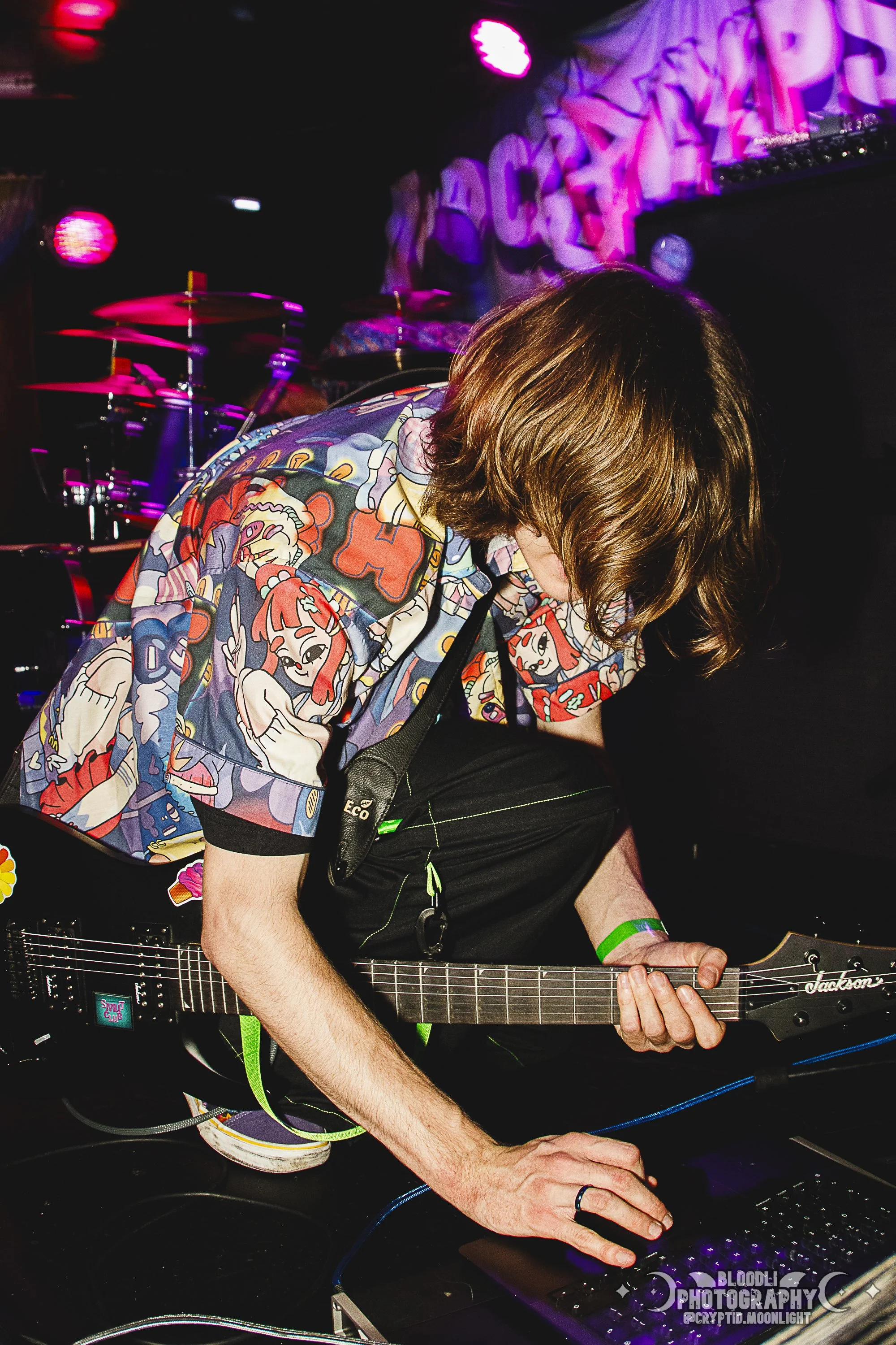 A person with shoulder-length brown hair wearing a colorful cartoon print shirt playing an electric guitar on stage, with a drum set and purple lights in the background.