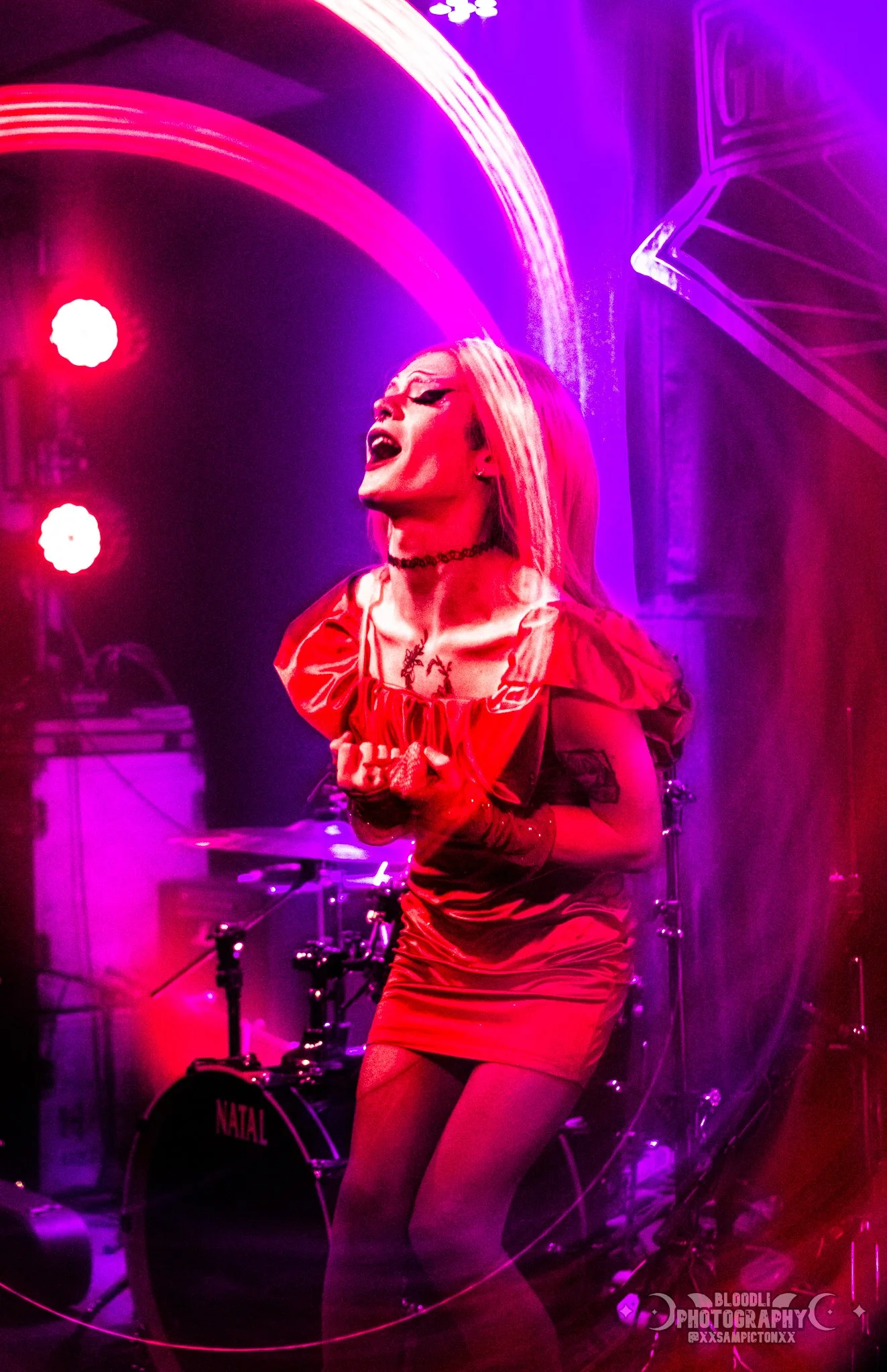 A female singer passionately performing on stage, with colorful purple and red stage lighting and a drum set in the background.