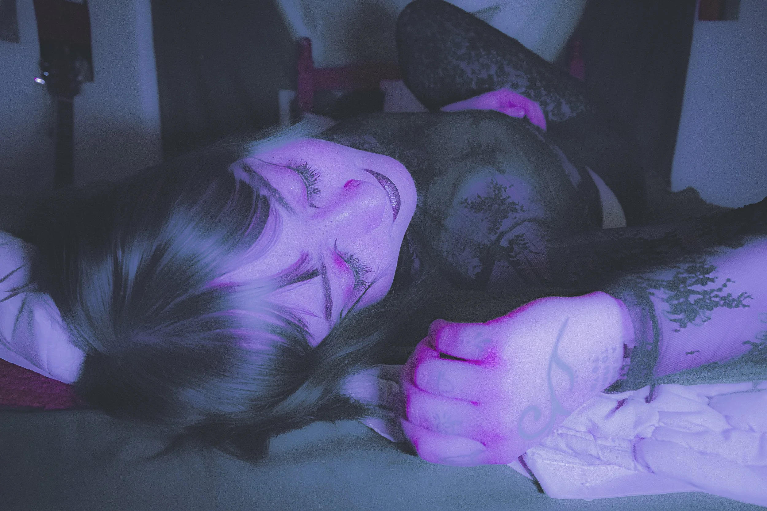 A woman lying on a bed with purple and pink lighting, smiling with closed eyes, wearing a black lace top, with tattoos on her hand, in a dimly lit room.