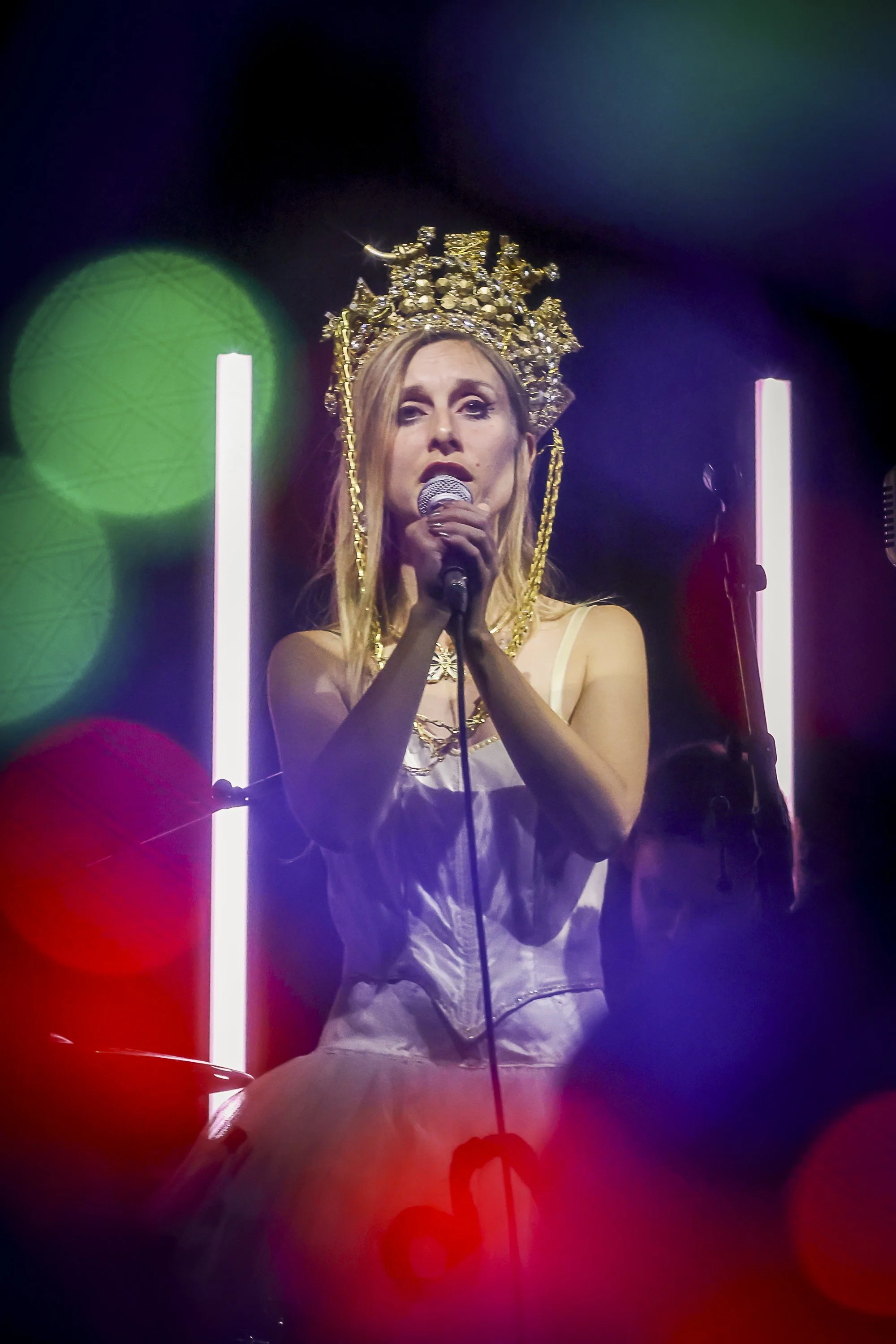 A woman wearing a gold crown and white dress, holding a microphone and singing on stage with colorful lights in the background.