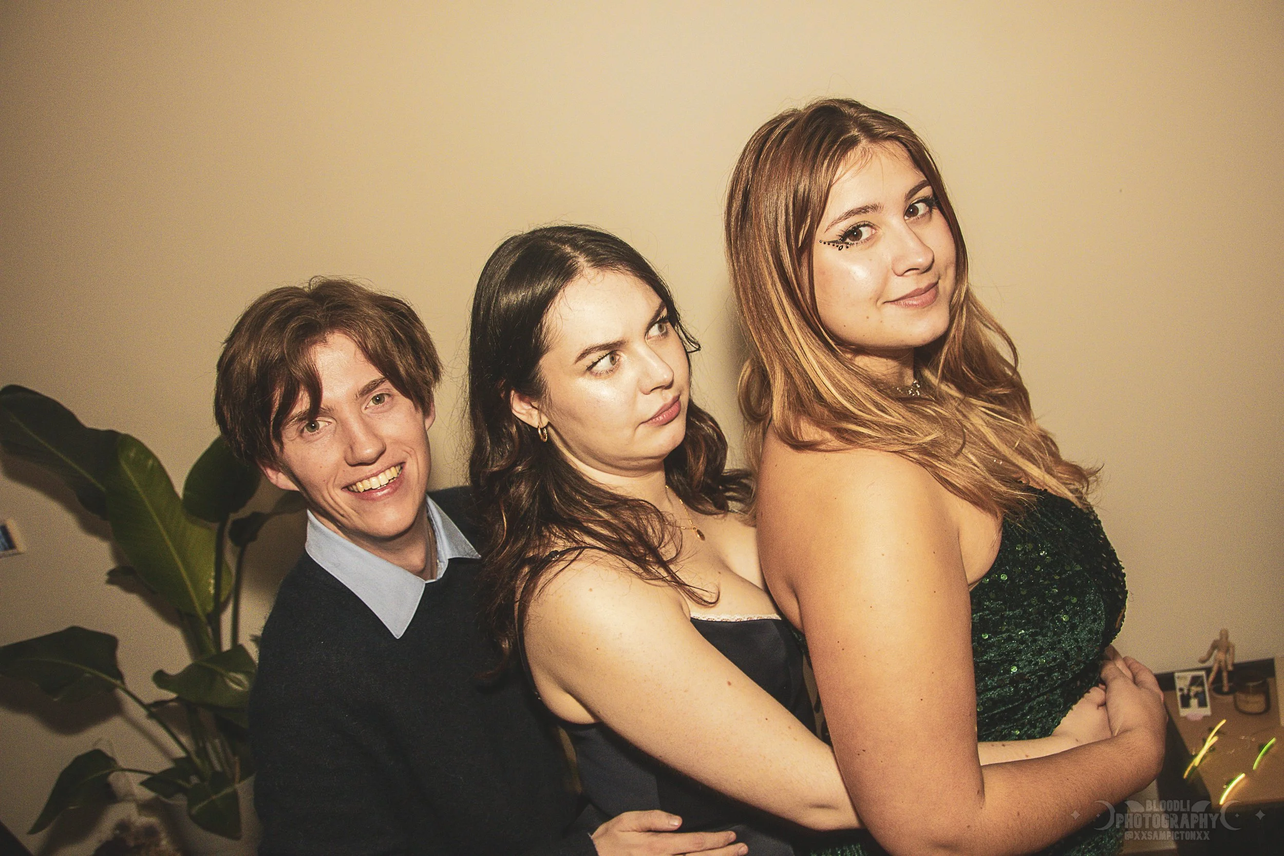Three young adults posing together indoors, with a large leafy plant in the background, smiling and looking at the camera.