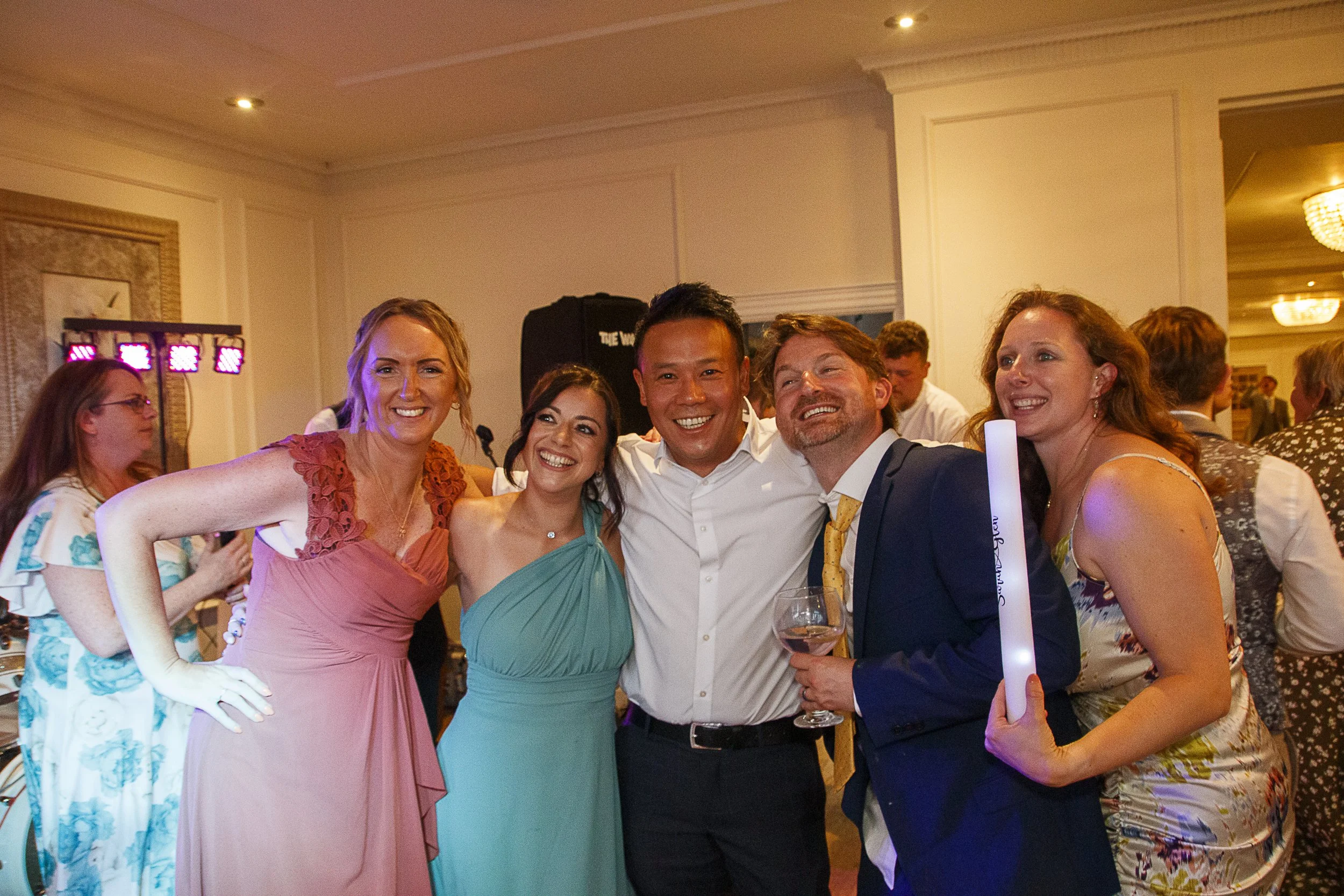 Six people in formal attire smiling at a celebration event, standing close together with arms around each other, holding drinks and a glow stick, in a decorated banquet hall.