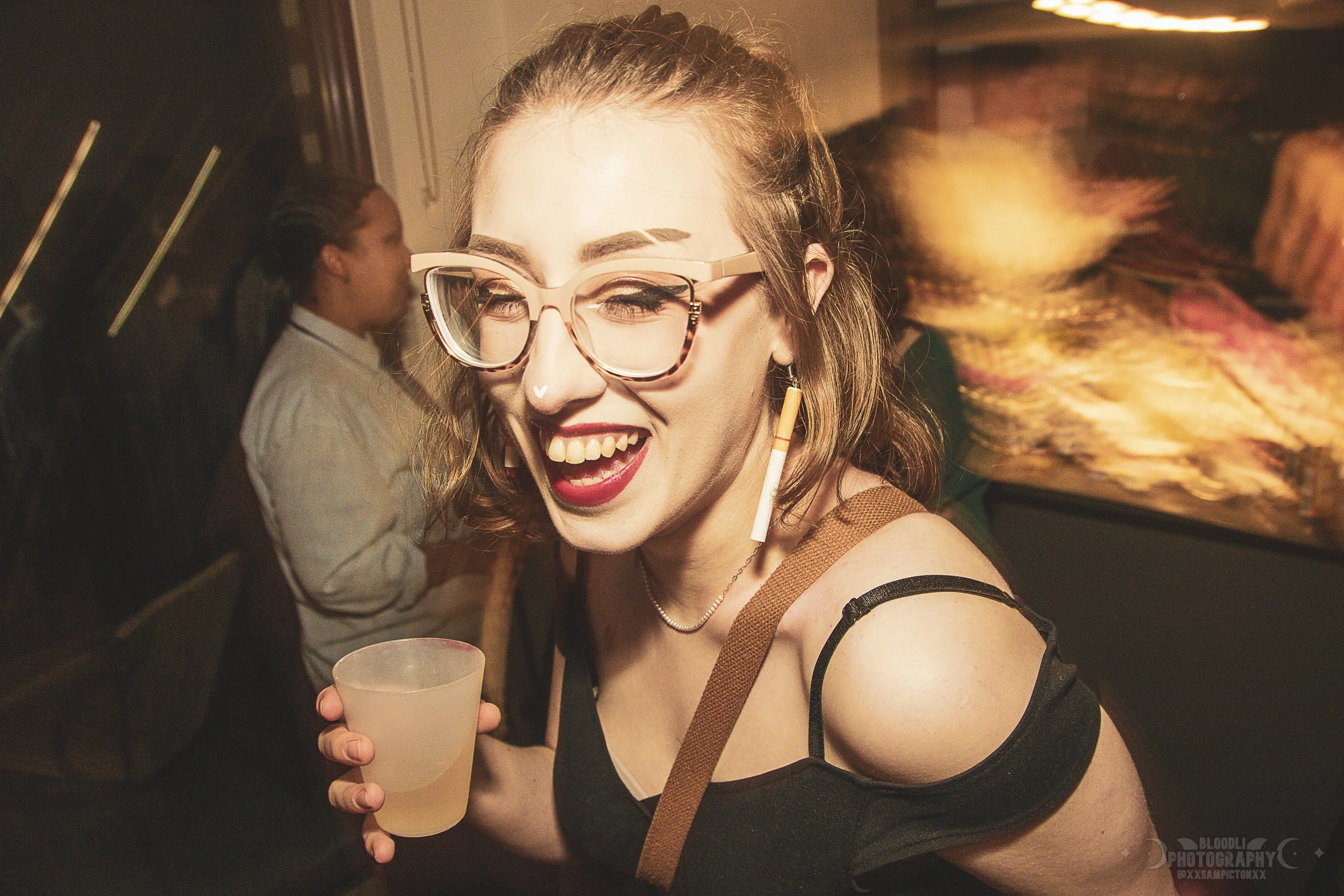 A young woman with glasses, lipstick, and a nose ring laughing at a party while holding a drink, with another person blurred in the background.