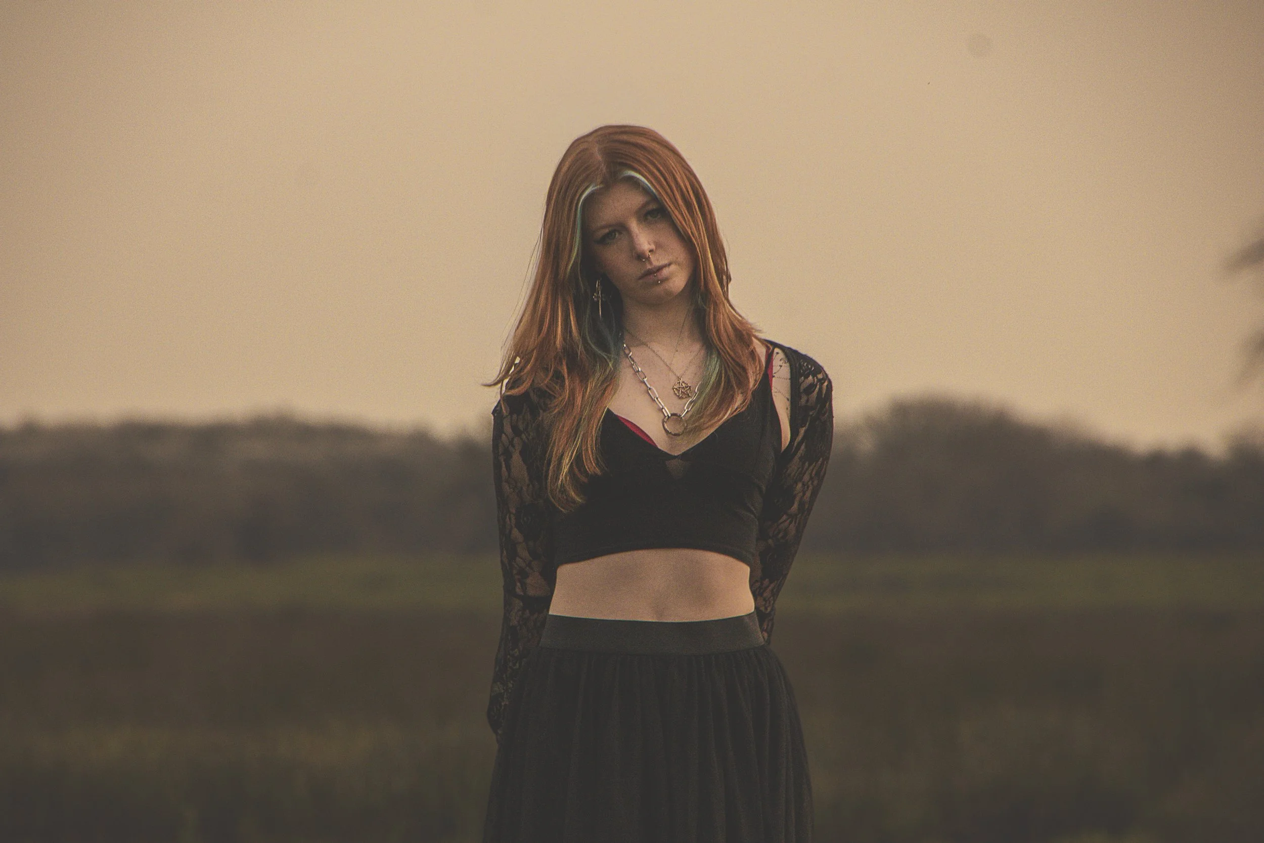 A young woman with long red hair stands outdoors during sunset wearing a black lace crop top and layered necklaces.