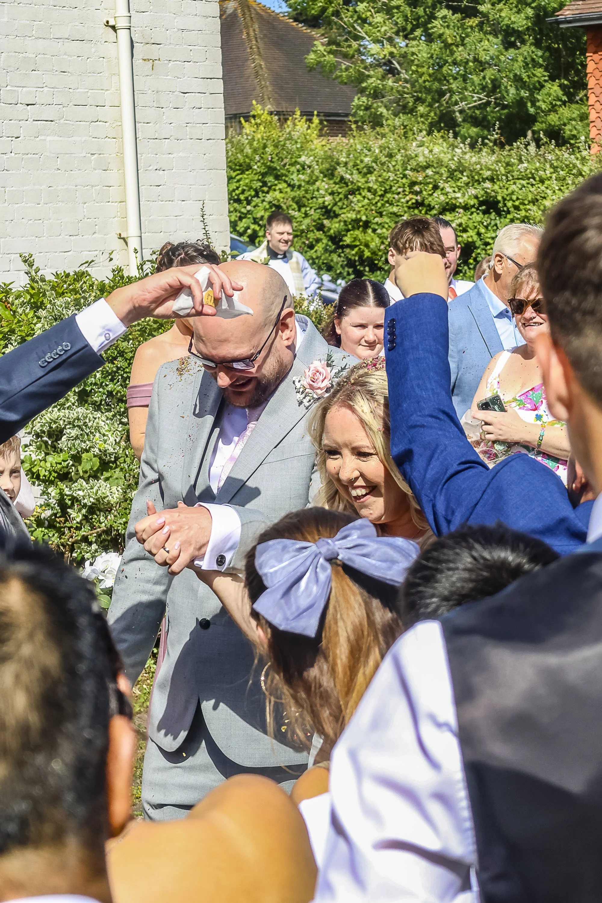People gathered outdoors during a celebration, some are arm-wrestling, with trees and houses in the background.