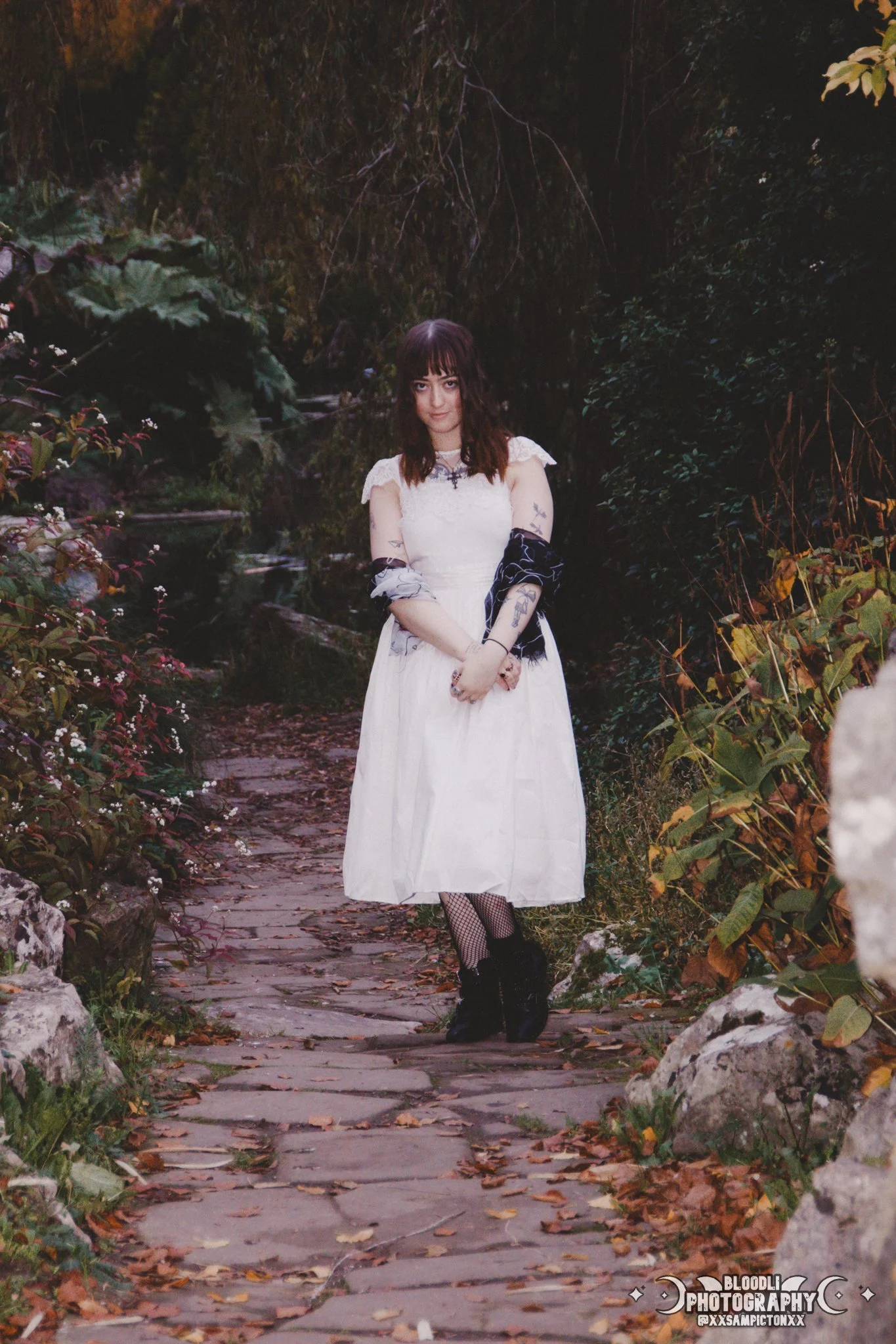 A young woman with tattoos, dark hair, and bangs standing on a stone path in a garden or forest area during daytime. She is wearing a white dress, fishnet stockings, and black shoes, with a dark shawl draped over her arms.