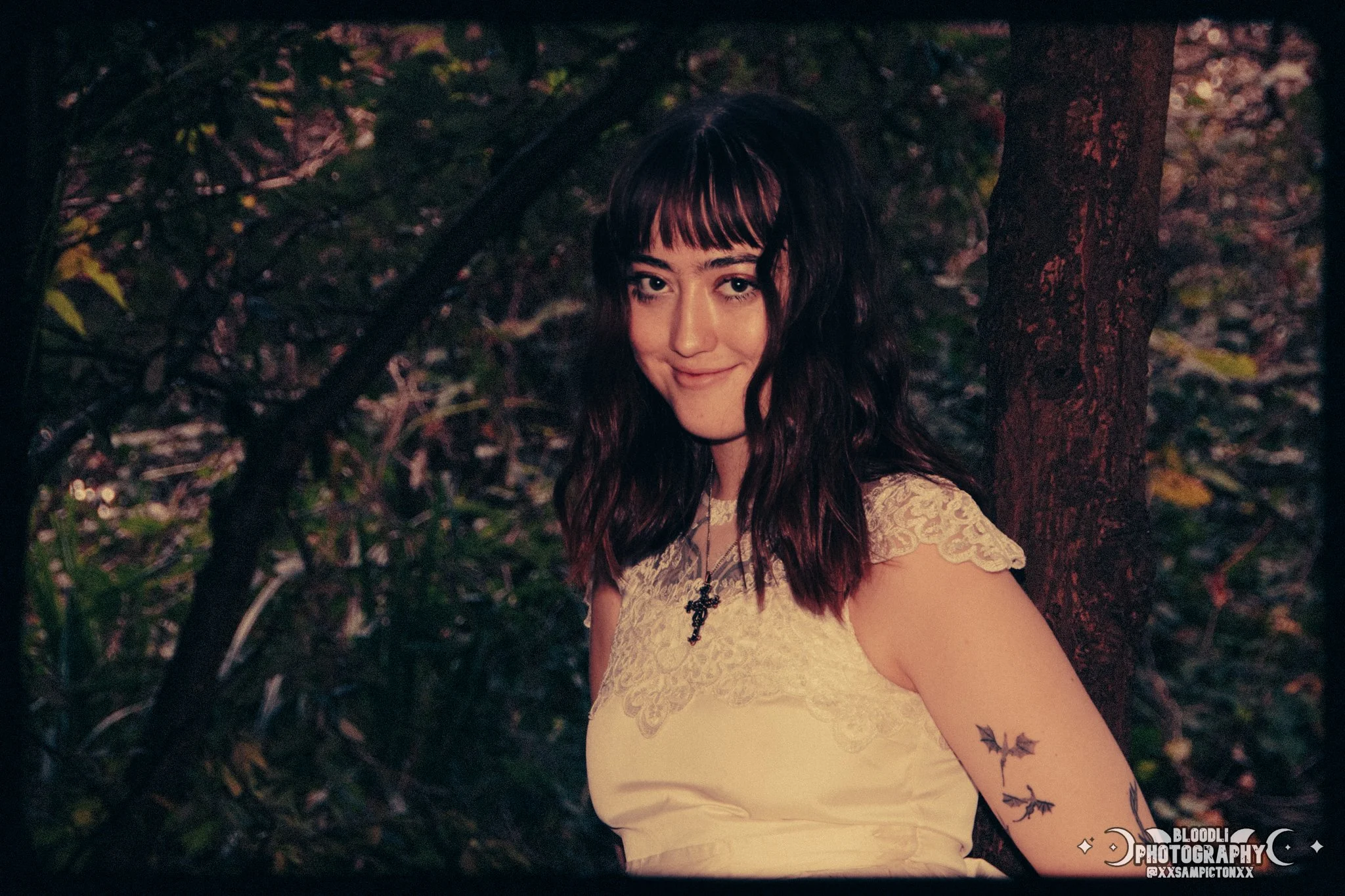 Young woman with dark wavy hair, wearing a lace cream dress and a cross necklace, standing outdoors next to a tree in a wooded area, smiling at the camera.