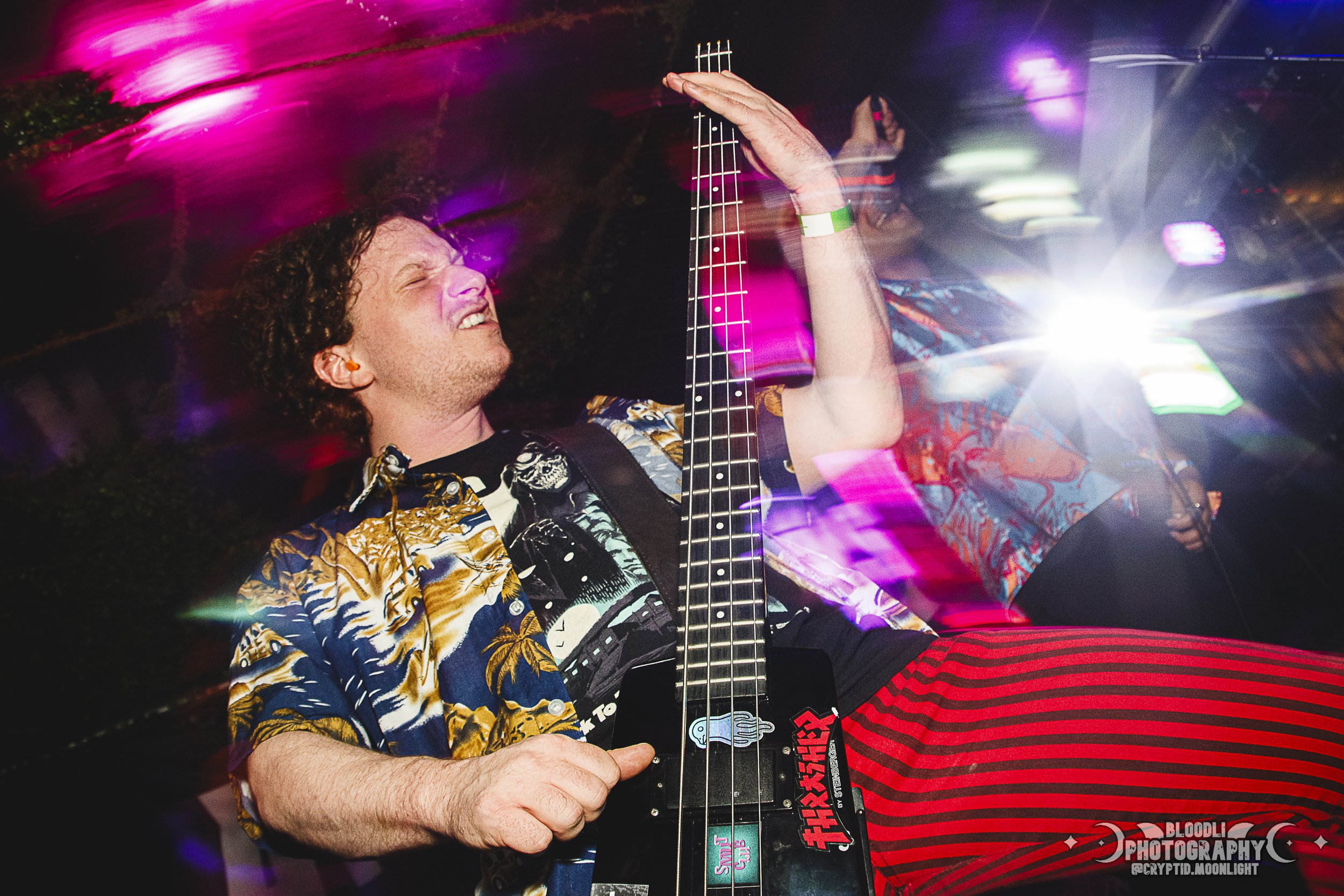 A man with curly hair playing an electric guitar at a lively party or concert, wearing a colorful Hawaiian shirt and striped red and black pants, with vibrant purple and pink streaks of light surrounding him.