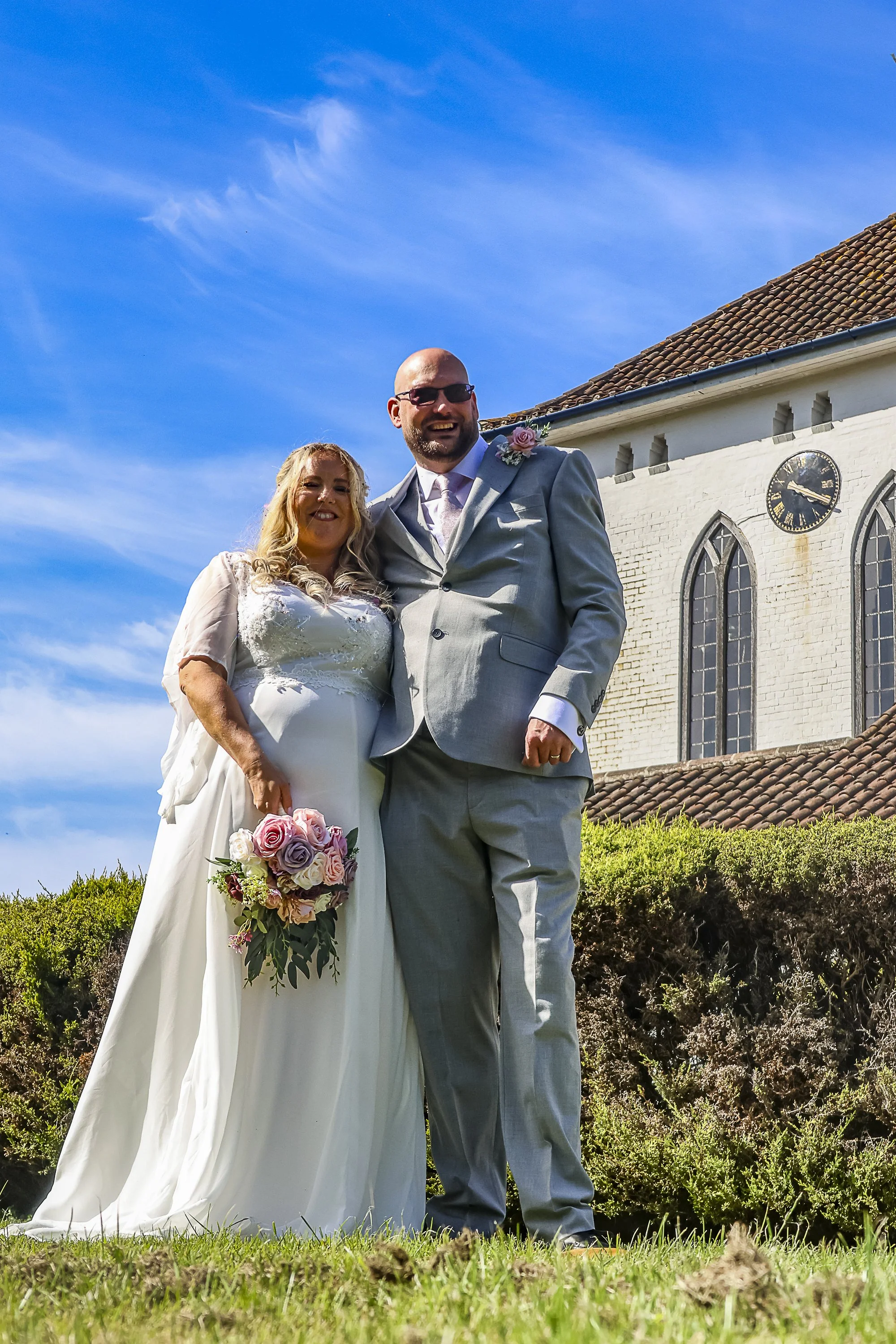 A bride and groom smiling and standing outside in front of a white church with a clock tower on a bright sunny day. The bride is holding a bouquet of pink, purple, and white roses, and wearing a white wedding dress. The groom is dressed in a light gr