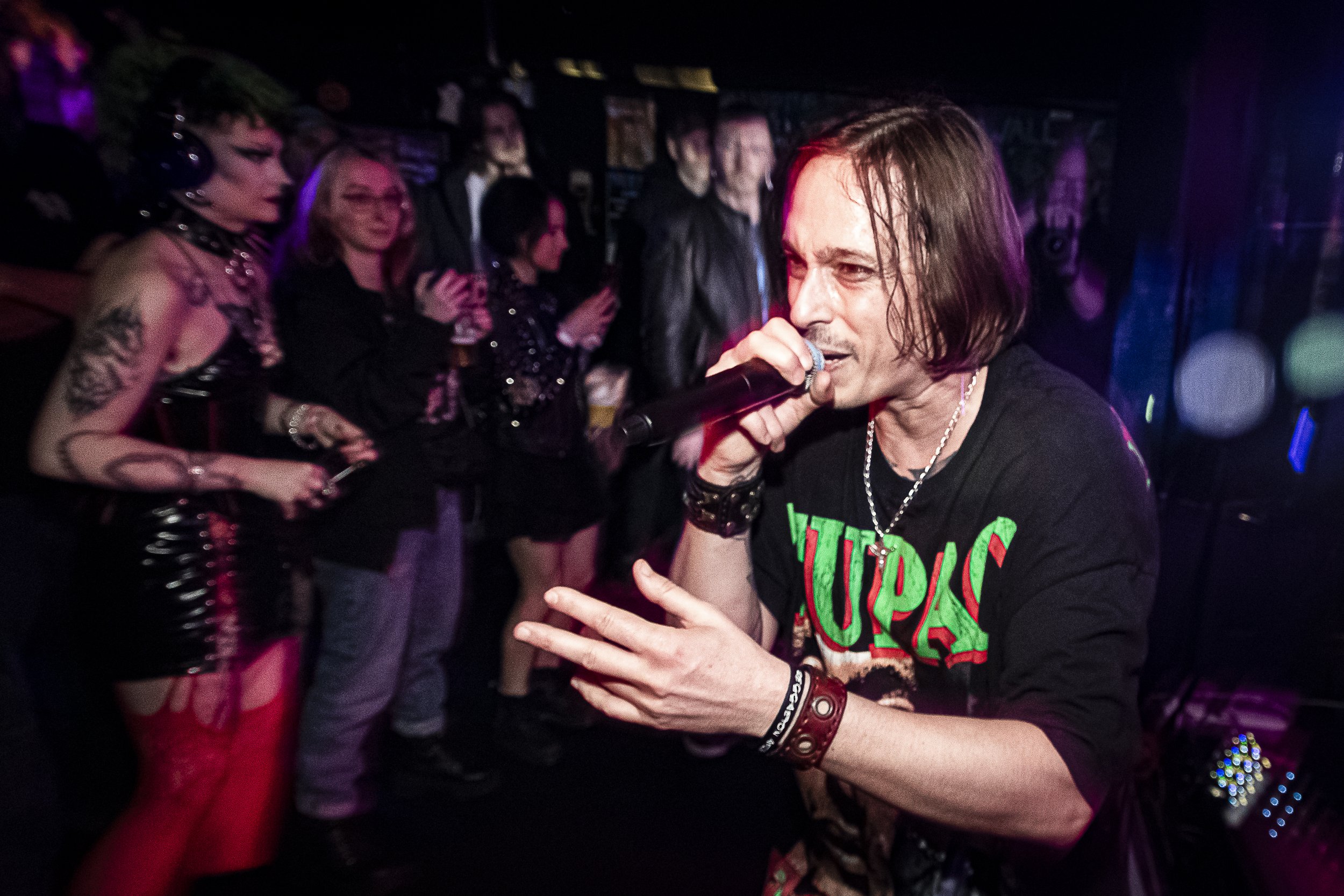 A male singer with shoulder-length hair performing with a microphone in a dimly lit venue, surrounded by an audience of women, some taking photos. The singer is wearing a black band shirt and various jewelry.