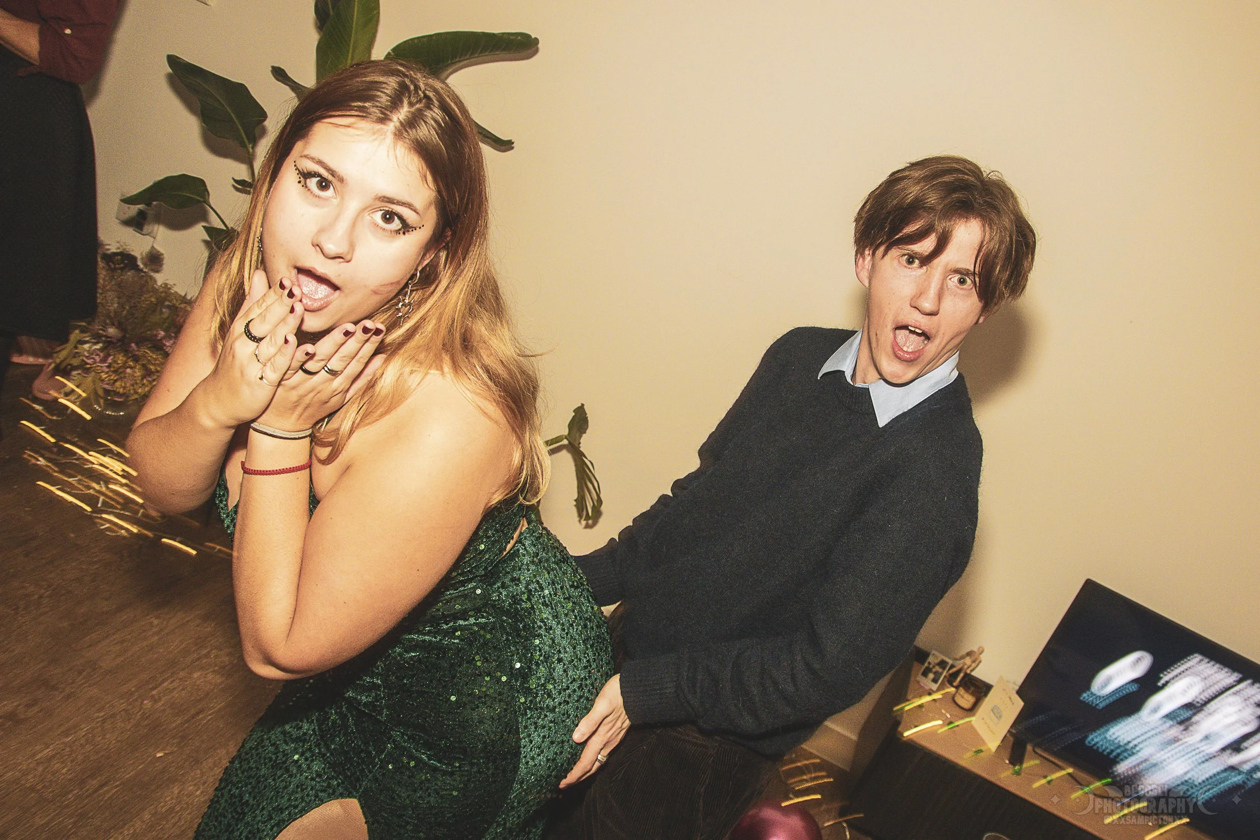 A young woman with long light brown hair, wearing a sparkly green dress, posing with her hand near her face at an indoor event. A young man with brown hair, wearing a dark sweater and a light blue collared shirt, is behind her with a surprised expres