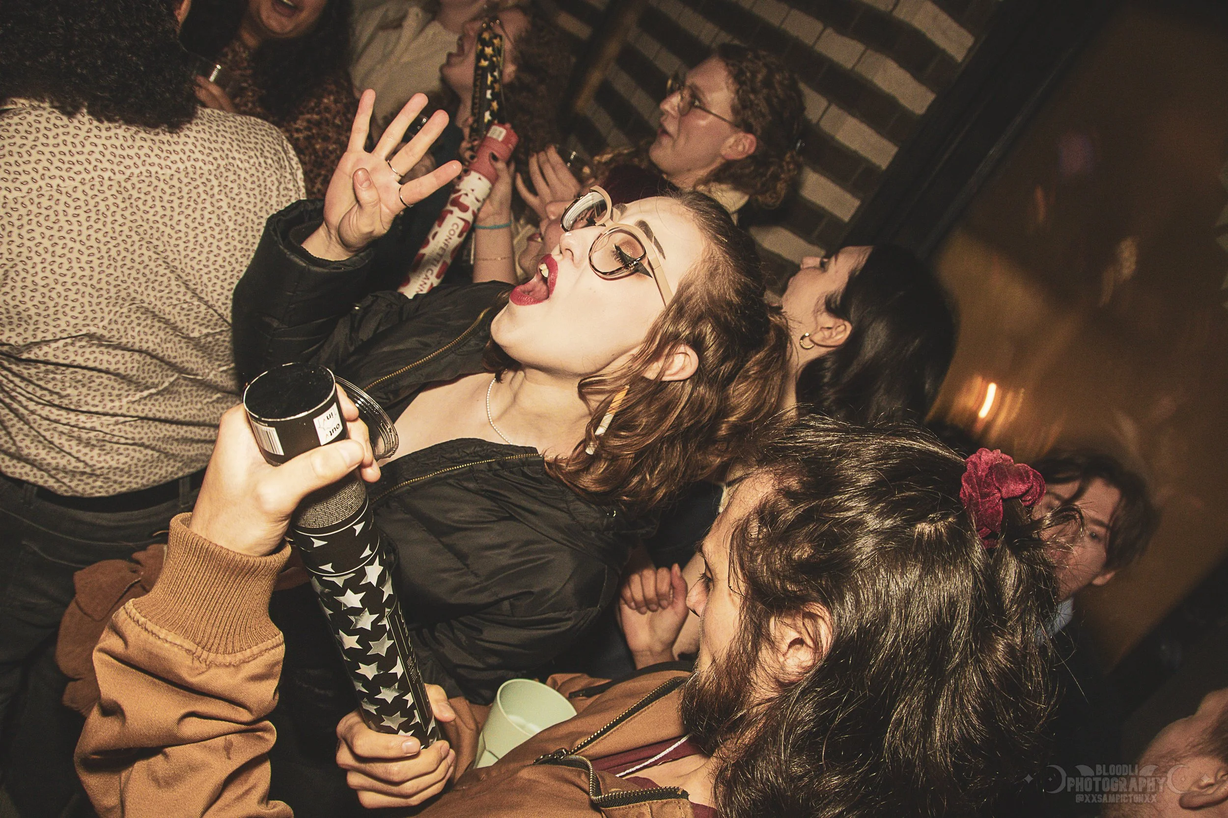 A group of young people at a party or club, with one woman in the foreground singing or speaking with her mouth open, wearing glasses and making a peace sign with her hand. Others around her are holding drinks and talking.
