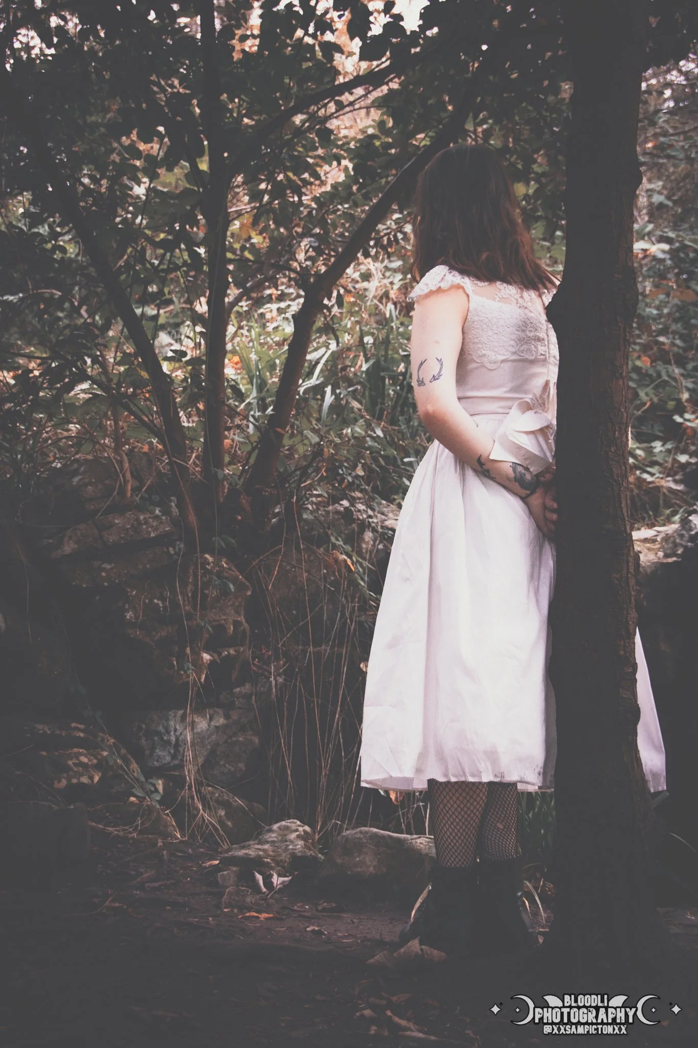 A woman standing outdoors among trees and rocks, wearing a white dress with lace details, fishnet stockings, and black boots, with tattoos visible on her arms.