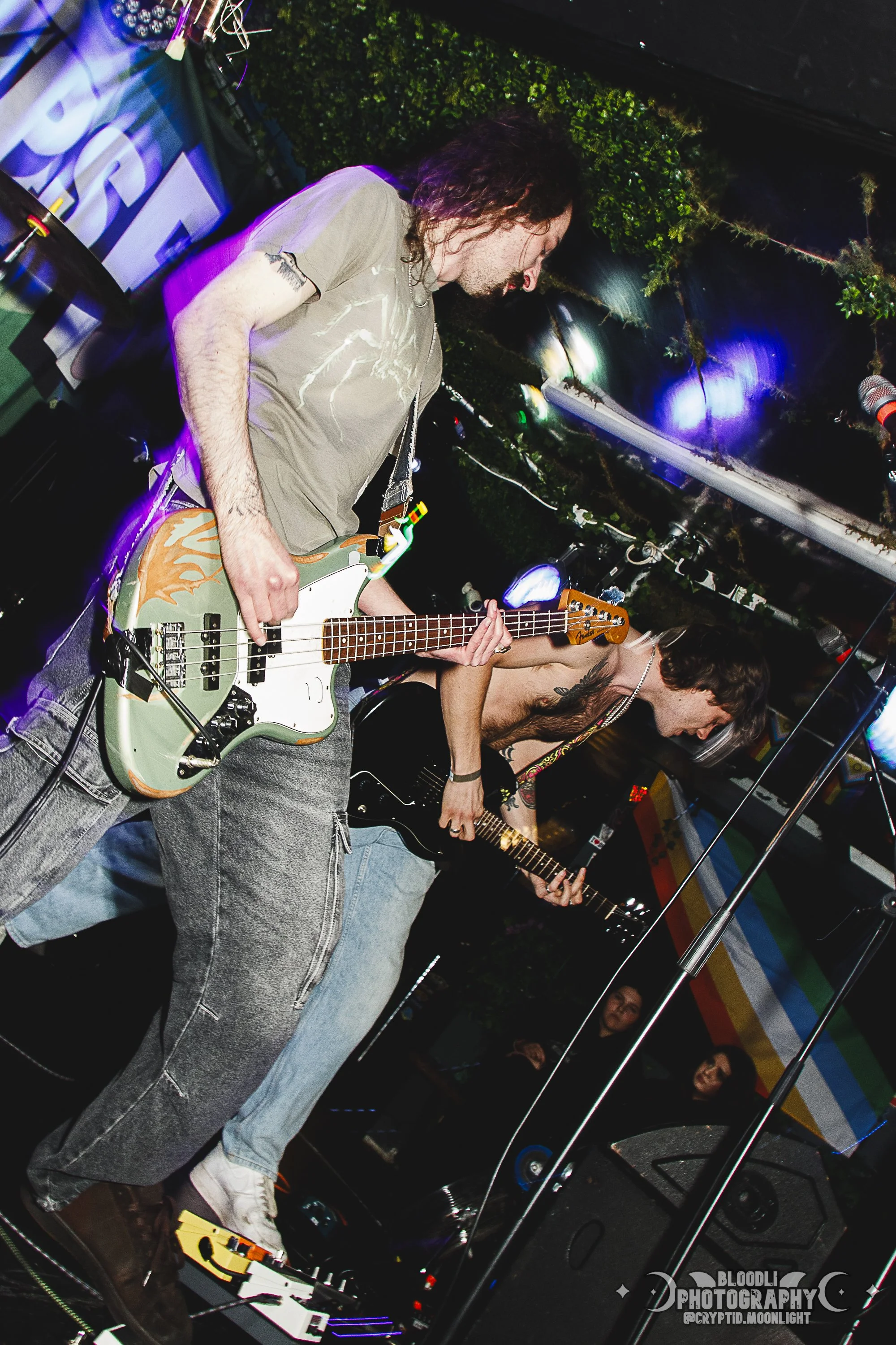 Two musicians playing guitar on stage during a live music performance, with a rainbow flag in the background and various stage equipment around them.
