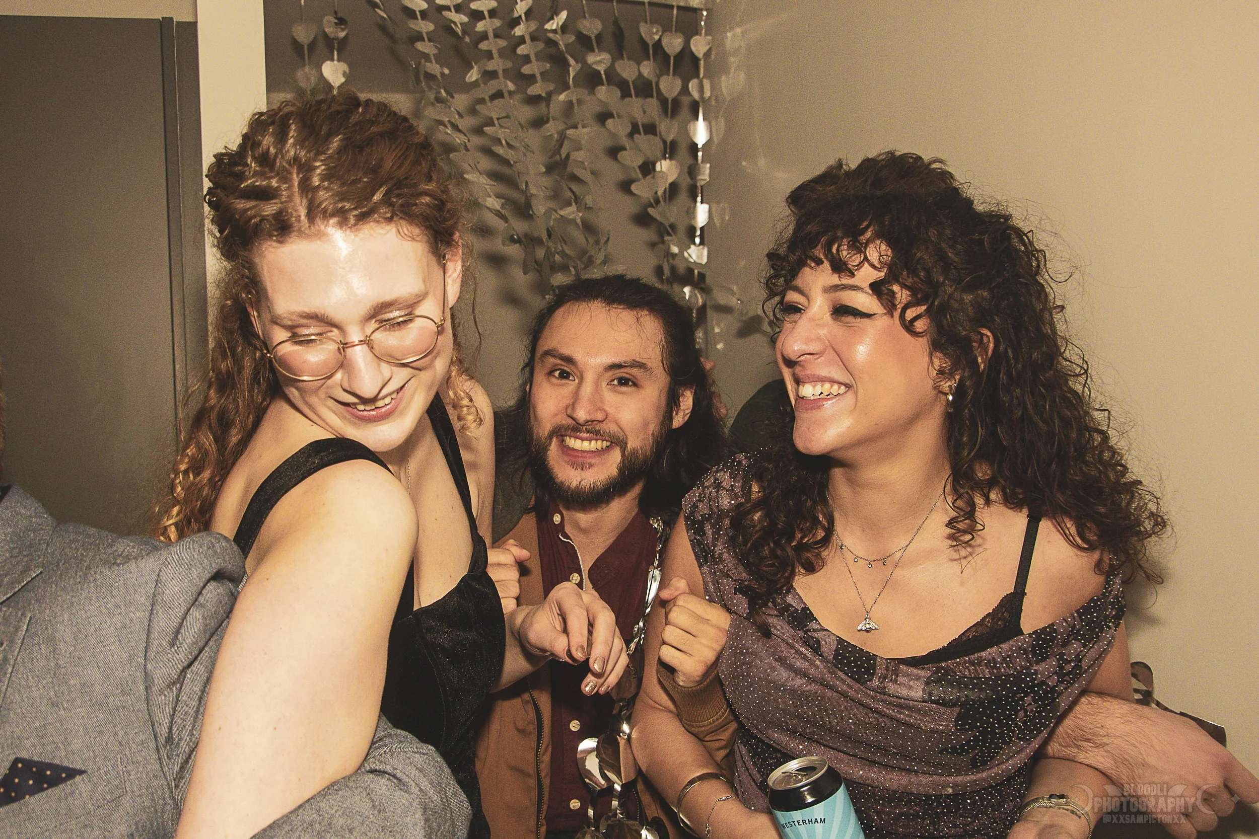 Three people smiling and laughing at a party, with a woman wearing glasses and a black dress on the left, a man with a beard and long hair in the middle, and a woman with curly hair holding a drink on the right.
