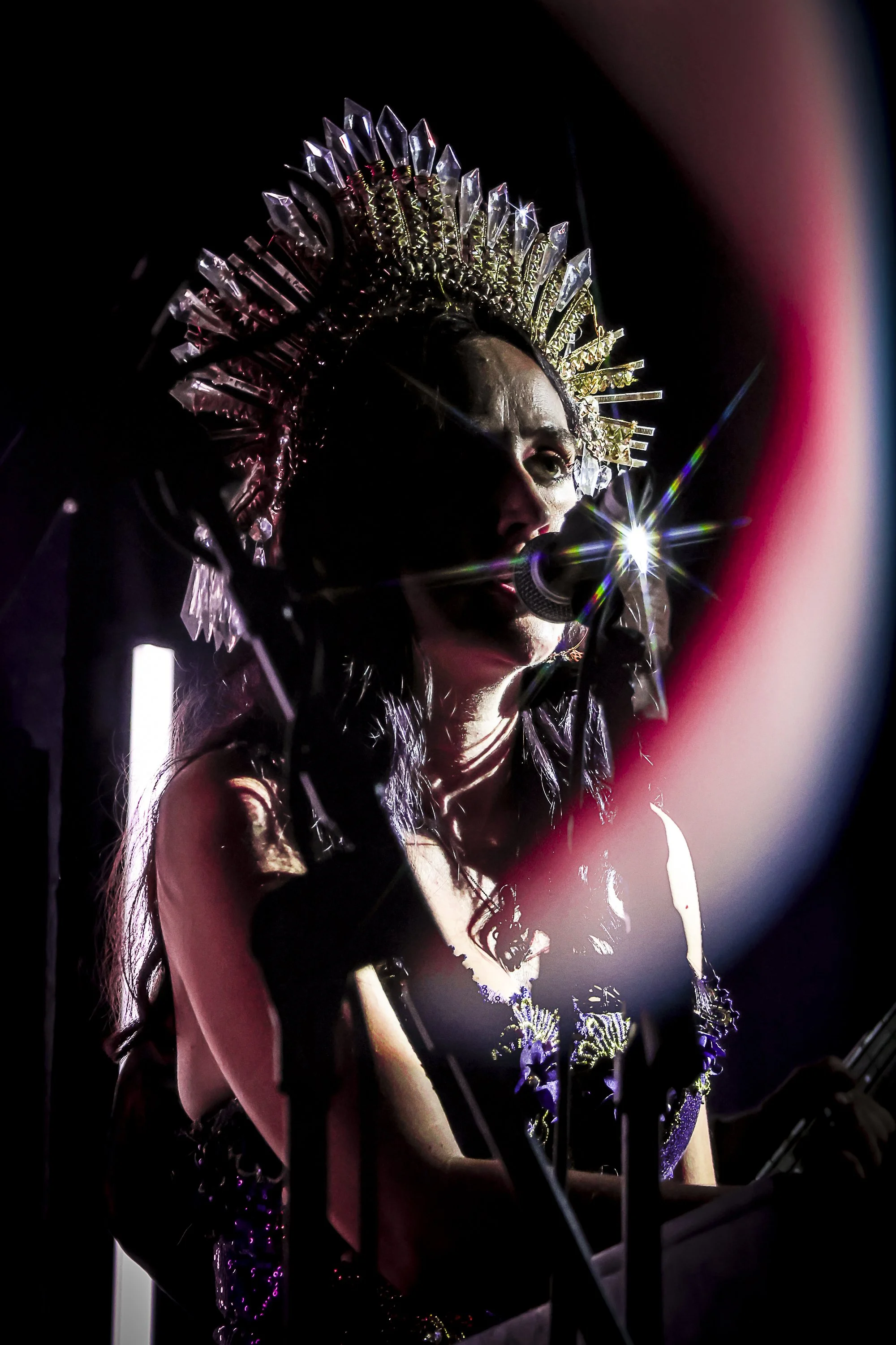 A performer wearing a large, ornate headpiece with crystals, singing into a microphone on stage, with dramatic lighting creating a starburst effect near her face.