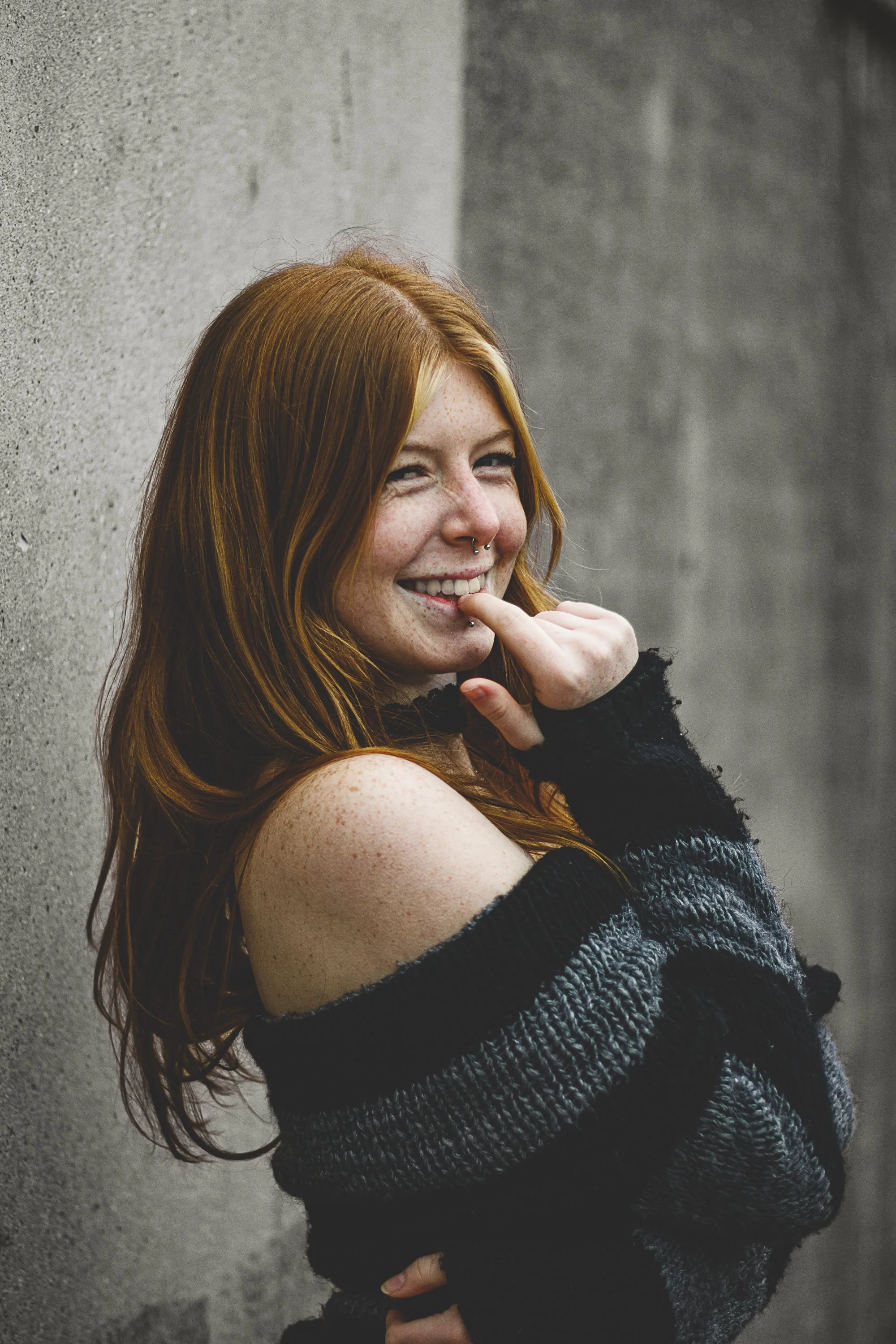 A smiling red-haired woman with a septum piercing, leaning against a concrete wall, wearing a black and gray off-shoulder sweater.