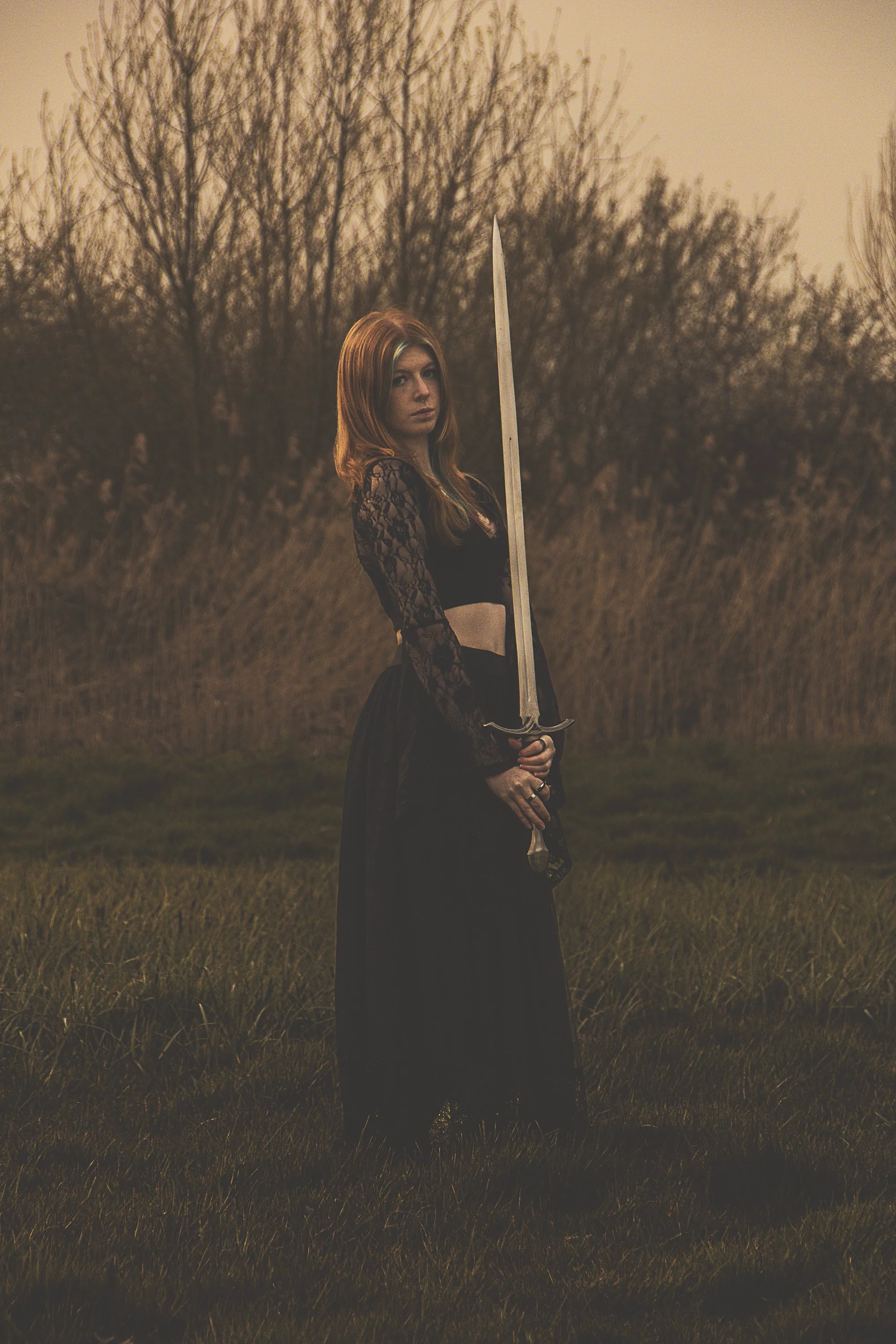 A young woman with long reddish hair standing outdoors in a field, holding a large sword vertically. She is wearing a black lace long-sleeve crop top and a long black skirt, with a serious expression as the sun sets behind leafless trees.