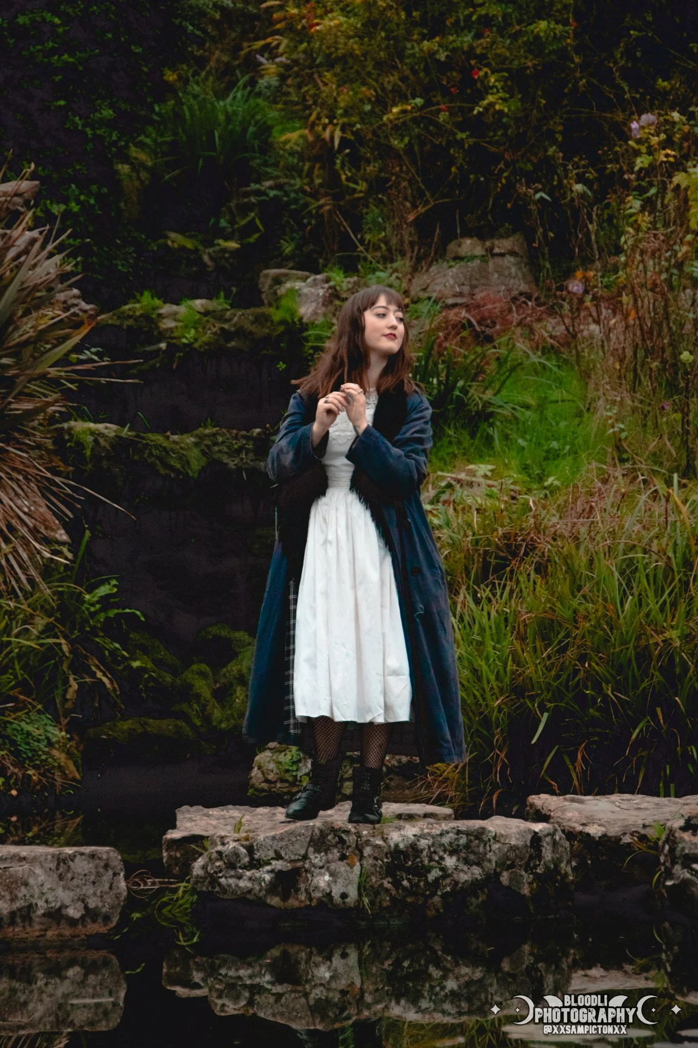 A young woman with dark hair and a white dress with a black coat standing on rocks by a small body of water in a lush, green garden.