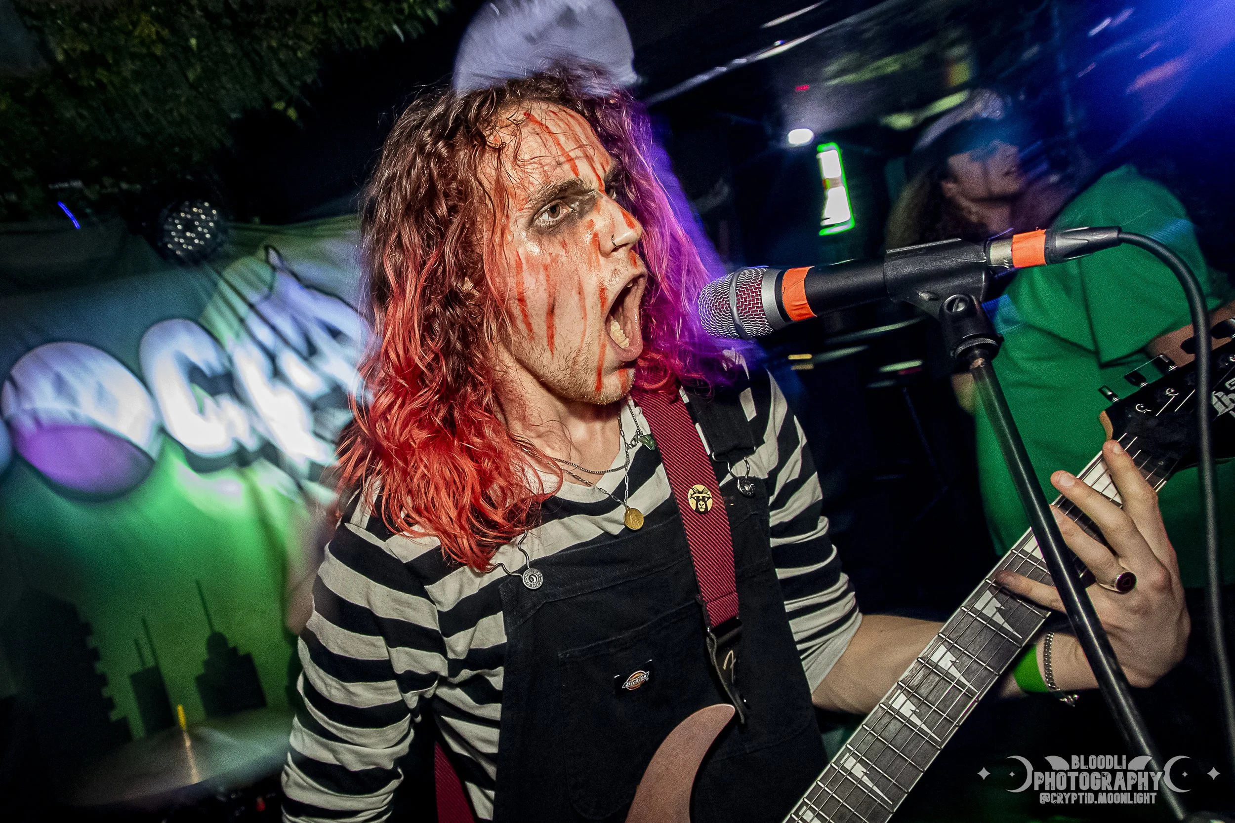 A person with long, curly hair painted with red streaks singing into a microphone while playing an electric guitar. They are wearing a black and white striped shirt and black overalls, in a lively indoor music scene.