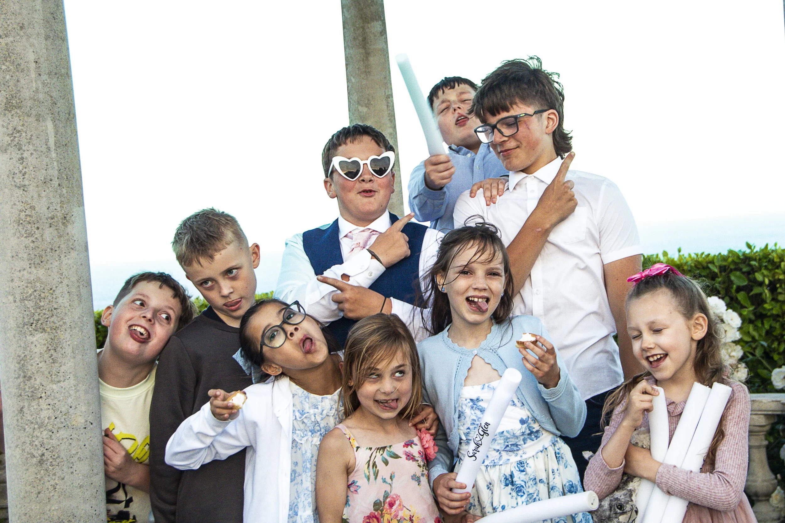 A group of children, some making funny faces and wearing sunglasses, gathered together outdoors during daytime. They are celebrating or attending an event.
