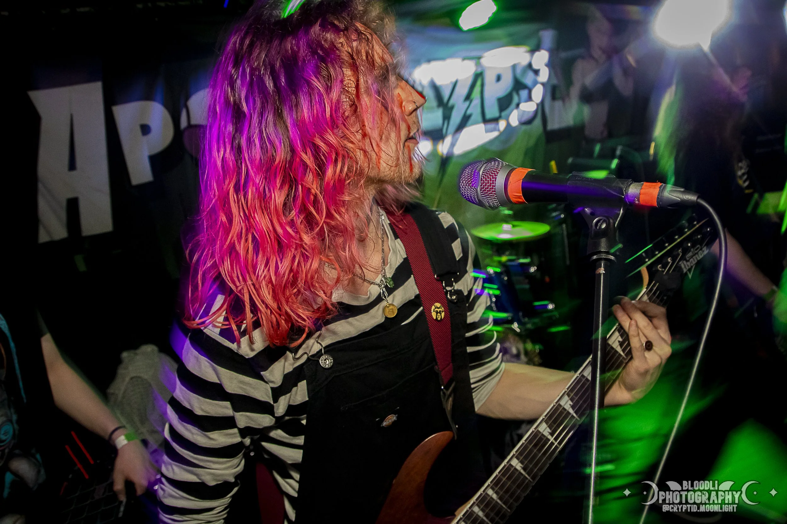 A woman with pink and red wavy hair wearing a black and white striped shirt, playing an electric guitar and singing into a microphone during a live music performance with colorful stage lighting.