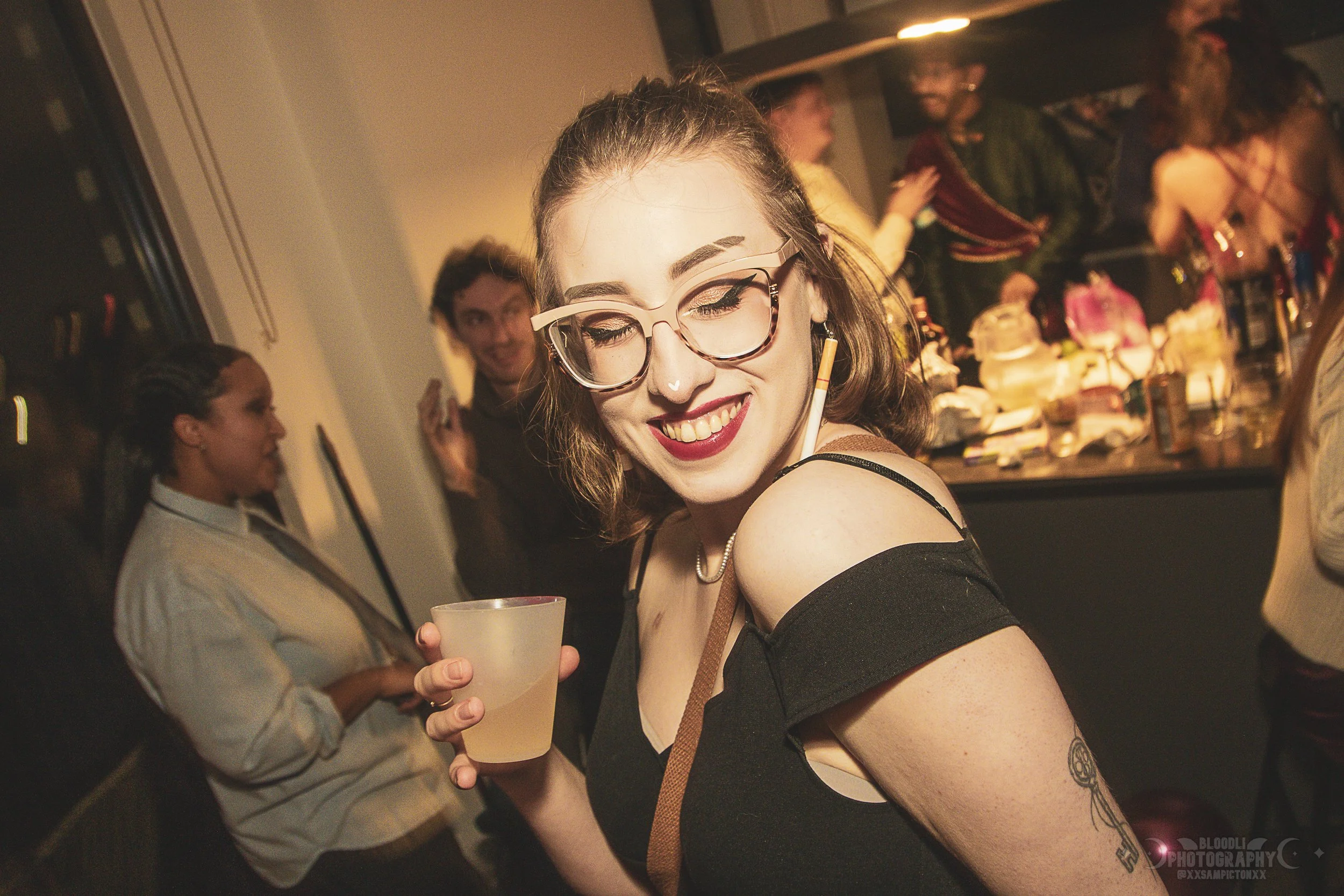 A young woman with glasses, makeup, and a nose piercing smiling and holding a drink at a party or celebration with other people in the background.