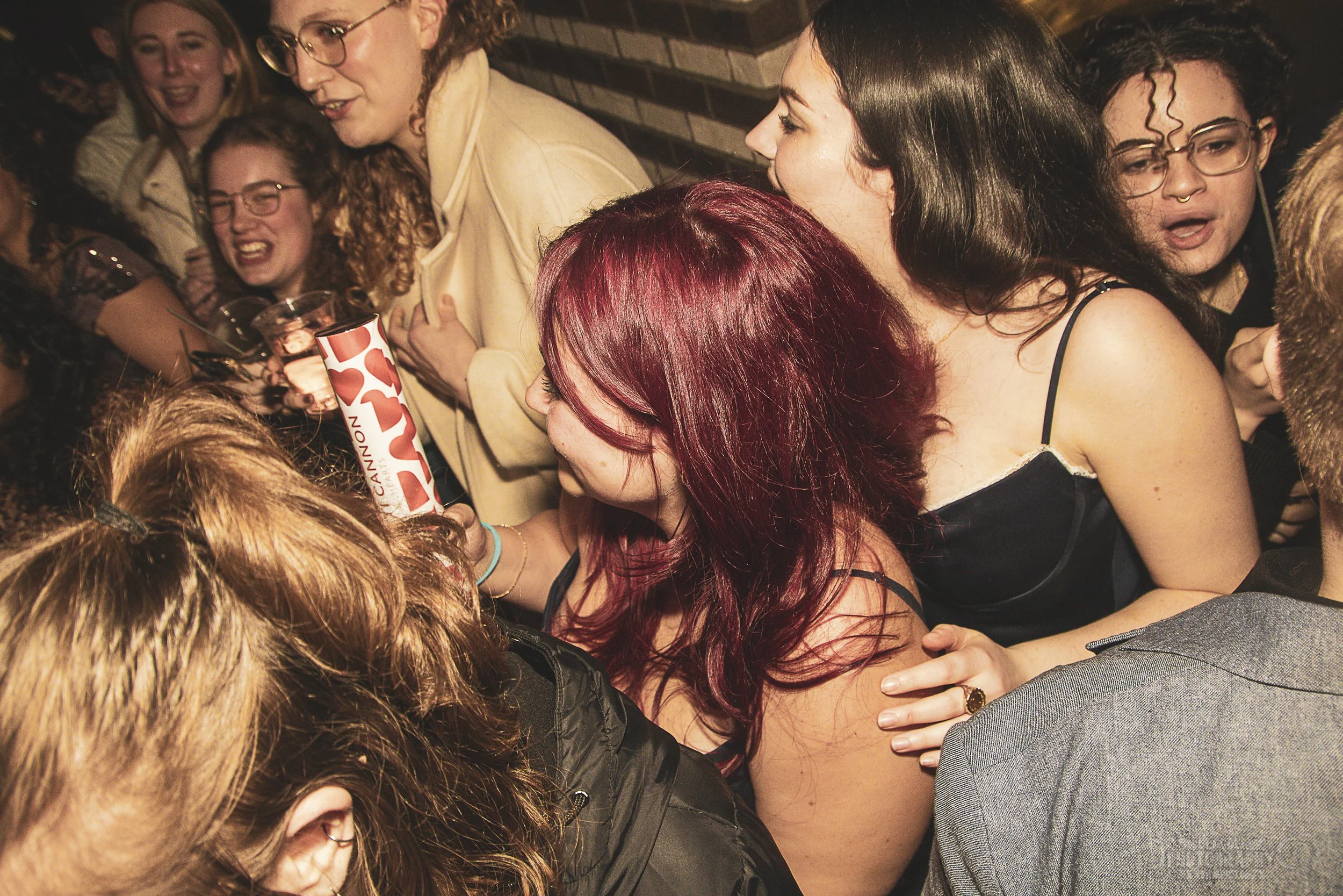 Group of people at a lively social gathering, laughing and dancing, with a woman holding a colorful drink in the center.
