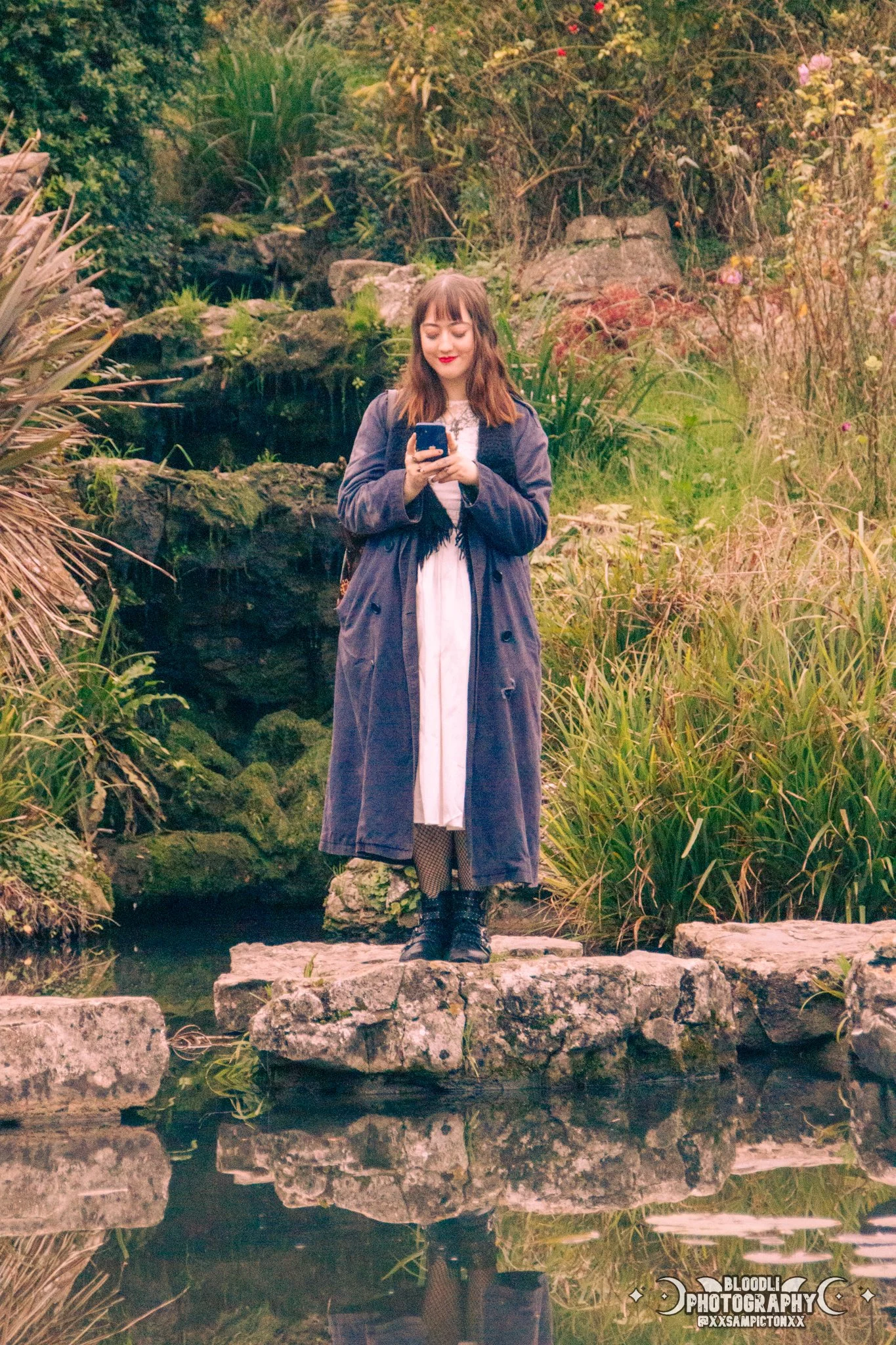 A young woman standing on rocks by a small pond, surrounded by lush greenery and plants, while looking at and smiling at her smartphone.