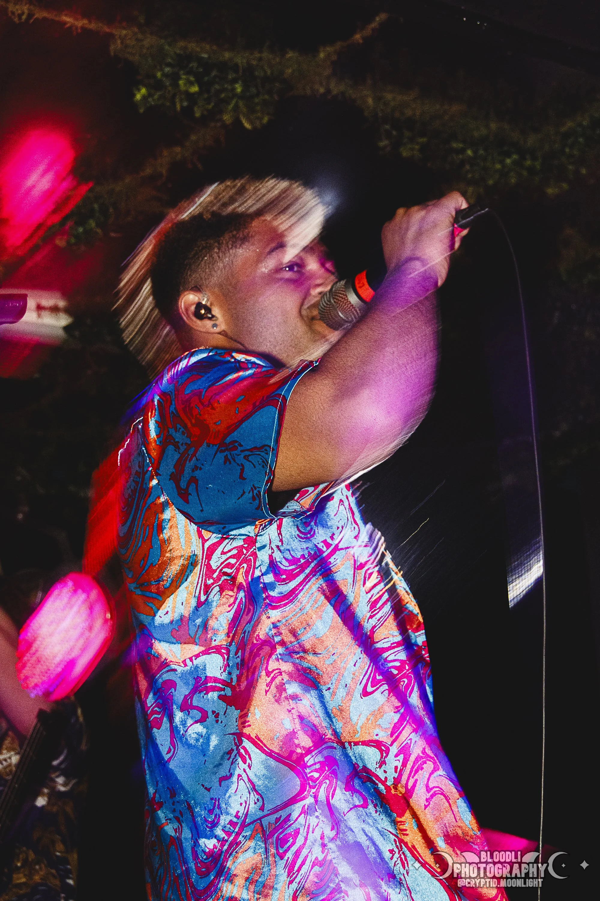 A person with blonde hair singing into a microphone during a live performance, wearing a colorful patterned shirt, with blurred movement and colorful stage lighting.