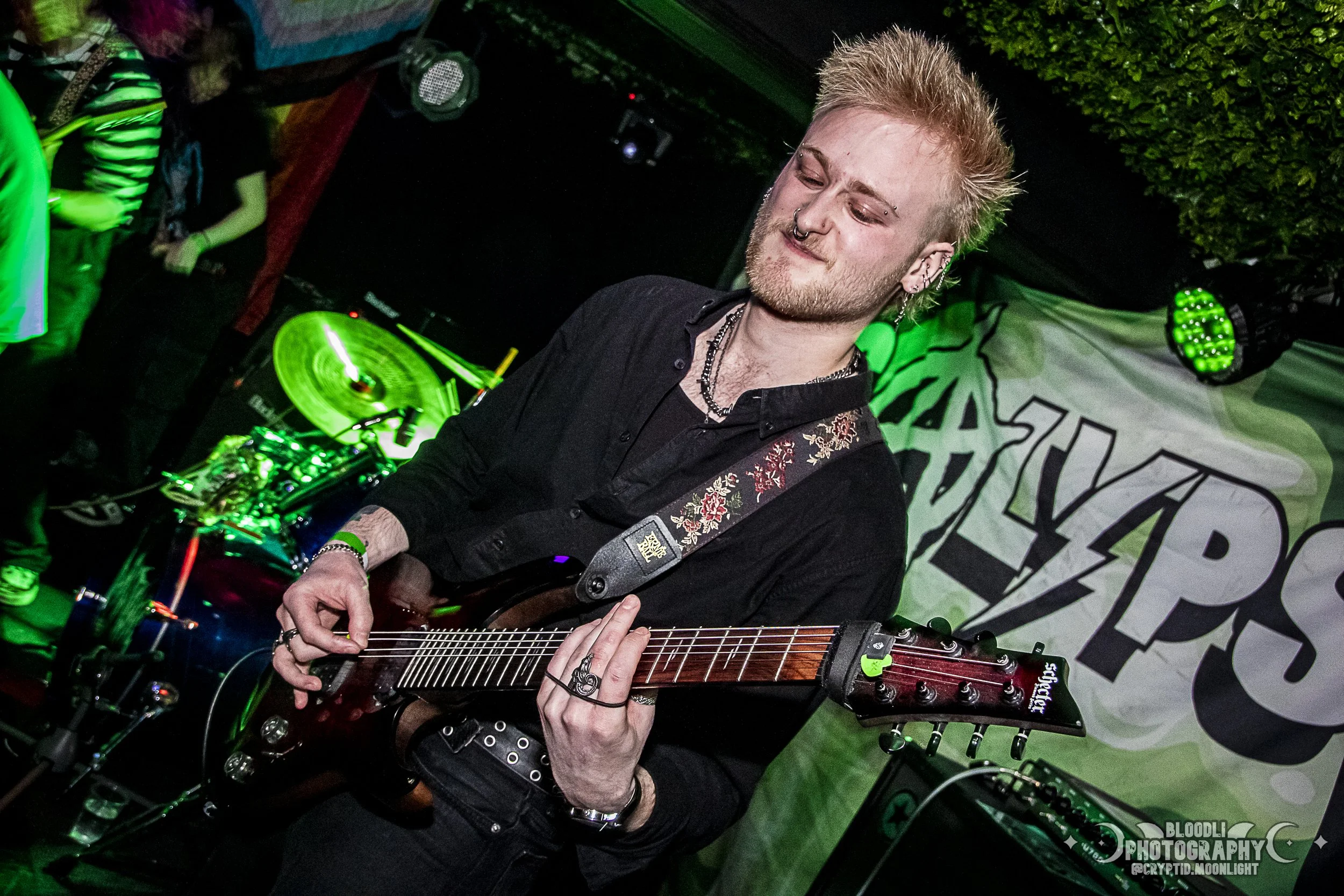Young man playing an electric guitar on stage with green lights and a Bloodless banner in background.