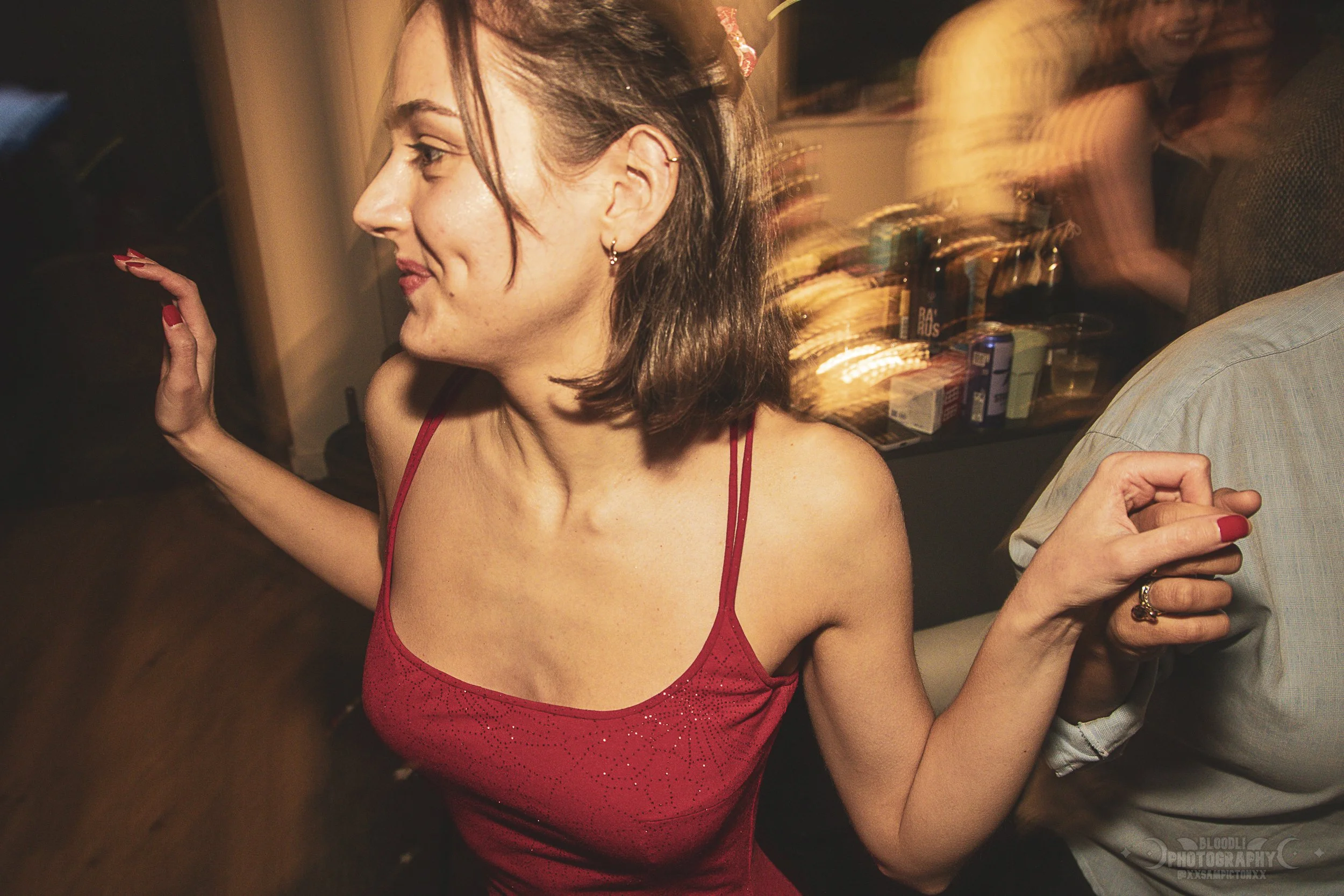 A woman in a red dress dancing at a party or social gathering, holding hands with someone, with blurred movement in the background indicating lively activity.