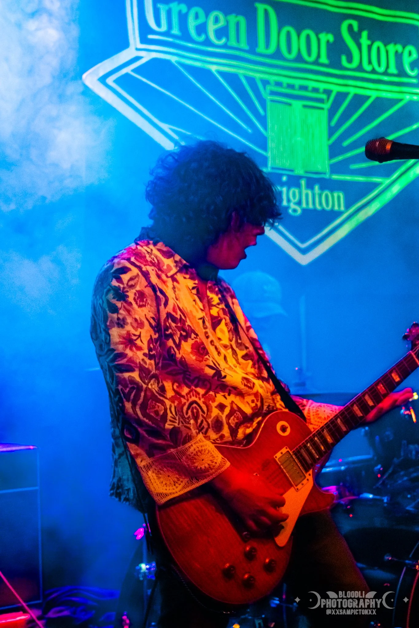 A person playing an electric guitar on stage with neon signs in the background, including a sign that reads 'Green Door Store'.