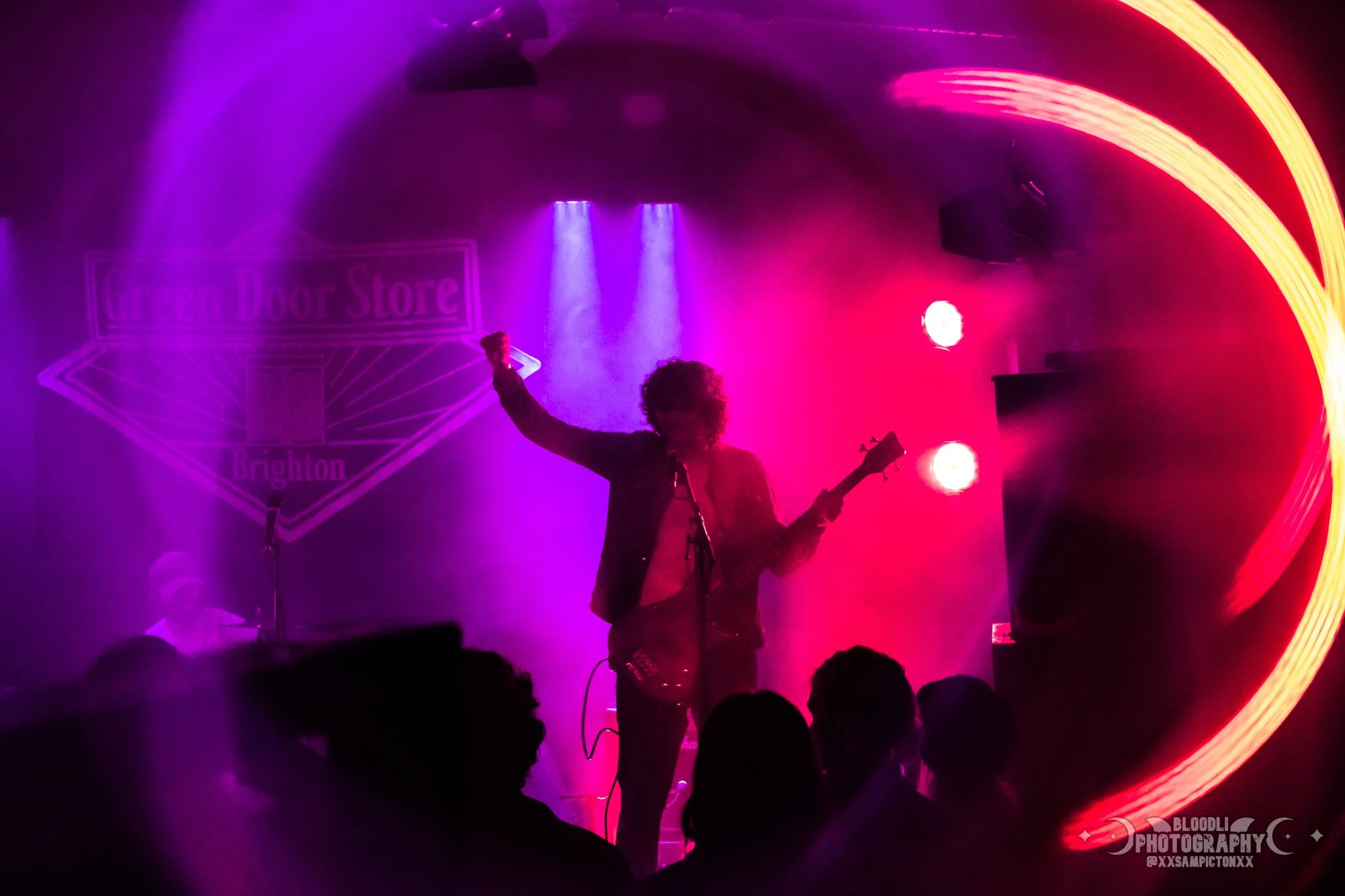 Silhouette of a musician with curly hair playing a guitar on stage, with colorful purple and pink stage lights creating a vibrant atmosphere. Audience silhouettes are visible in the foreground, and a neon sign reading 'Queen Door Store Brighton' is i