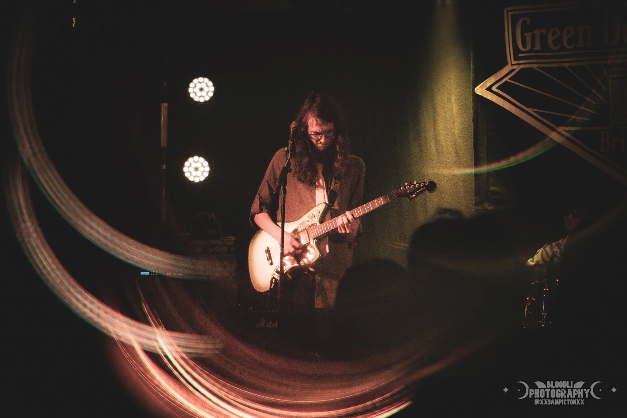 A person with long hair and glasses playing an electric guitar on stage with stage lights and a dark background.