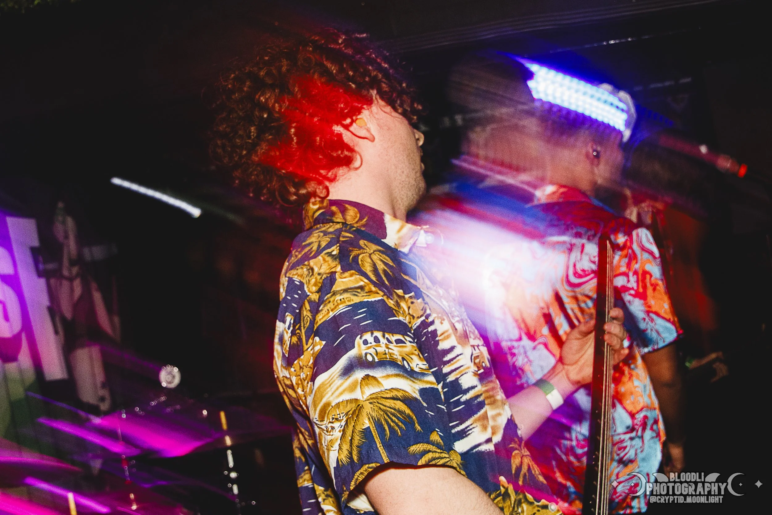 Two musicians perform at a nightclub, one playing the keyboard and the other holding a microphone. Both wear colorful, tropical-themed shirts, and vibrant lights illuminate the scene.