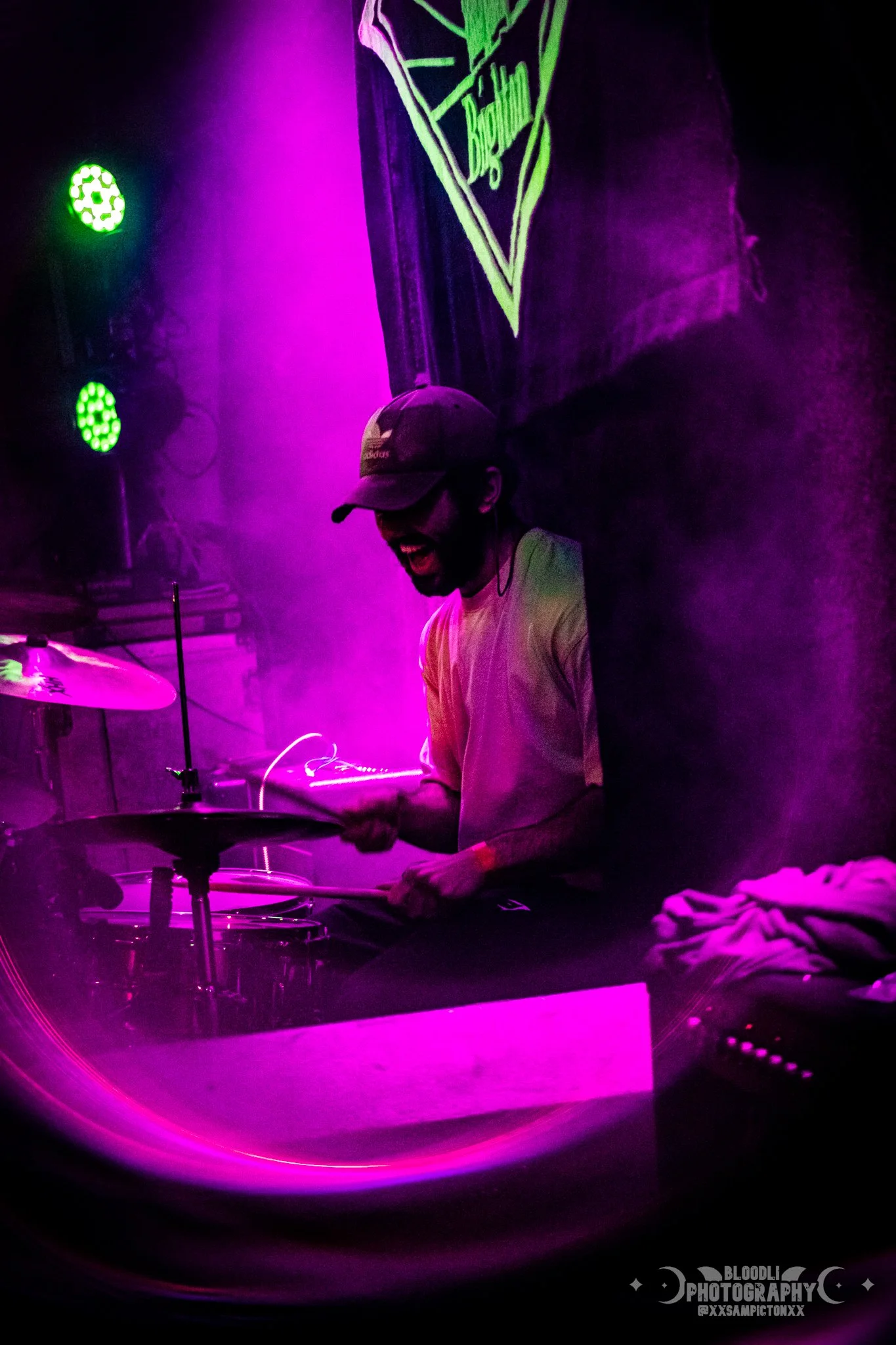 A person playing drums on stage with purple and green lights, wearing a gray cap and white shirt, smiling and enjoying music.