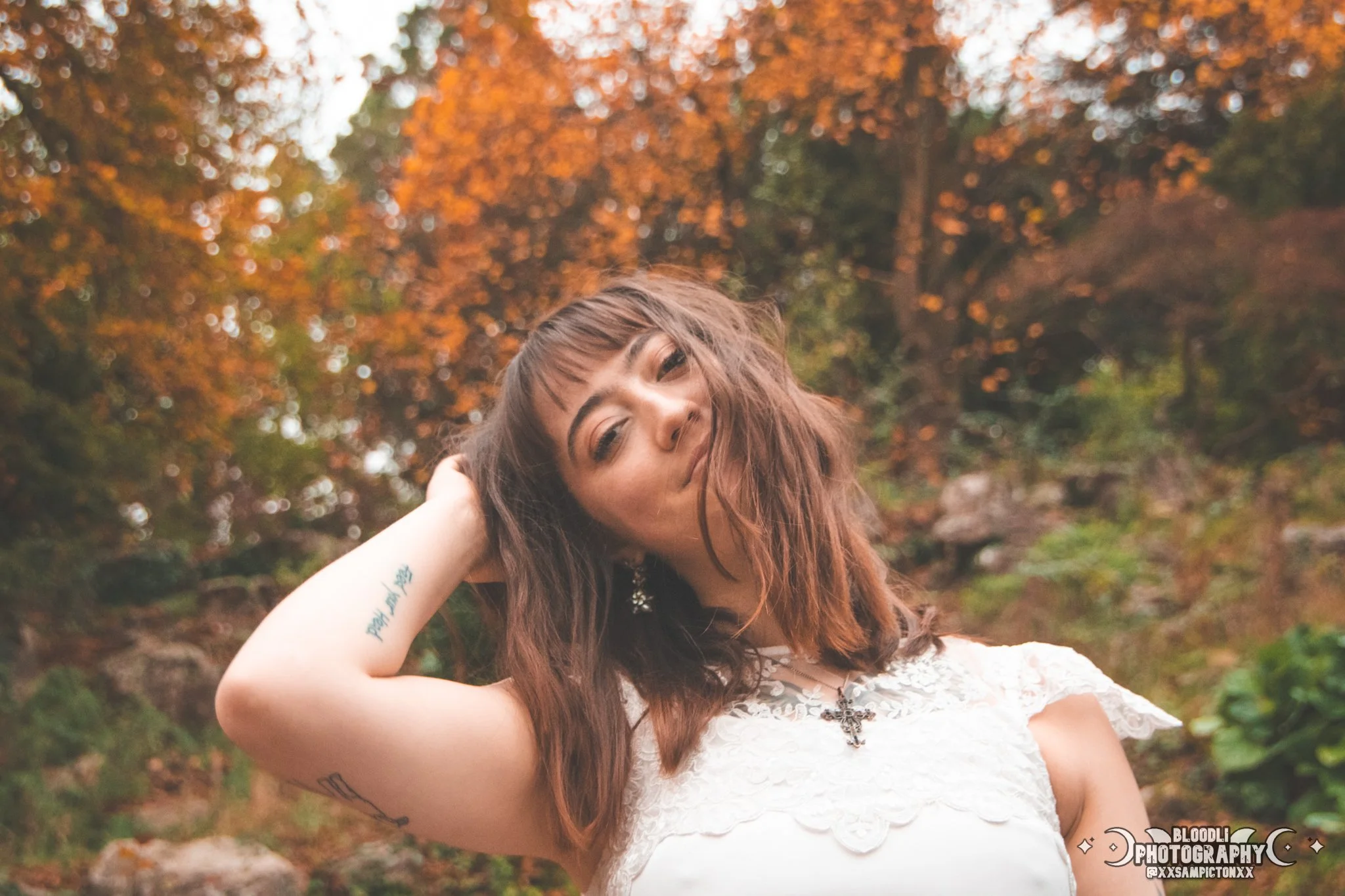 Young woman with brown hair and tattoos wearing a white lace dress outdoors in a fall forest with orange leaves.