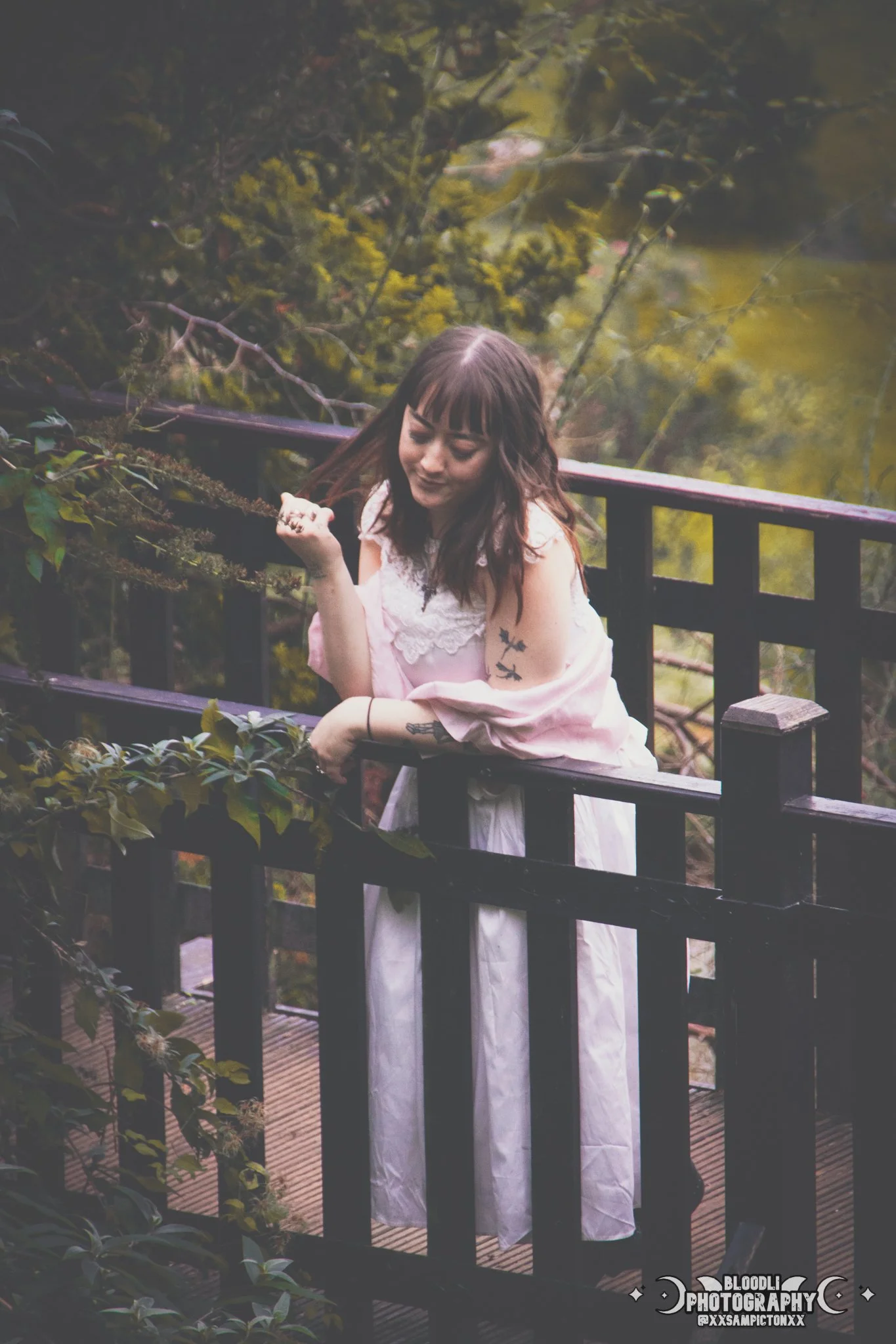 A woman with dark hair and bangs, wearing a pink dress, leaning on a black wooden railing on a wooden deck outdoors surrounded by lush greenery.