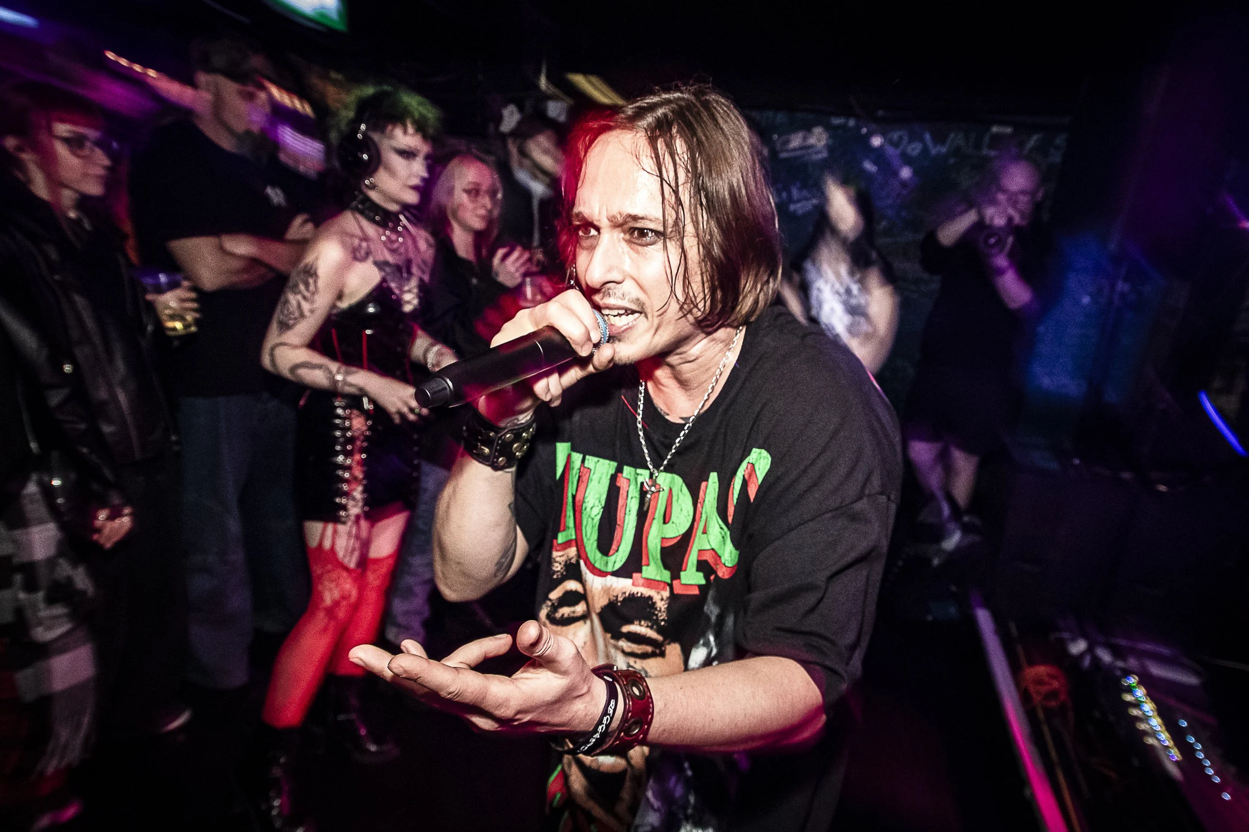 A man with shoulder-length hair singing into a microphone at a lively, crowded party or concert, with people in goth and punk fashion in the background.
