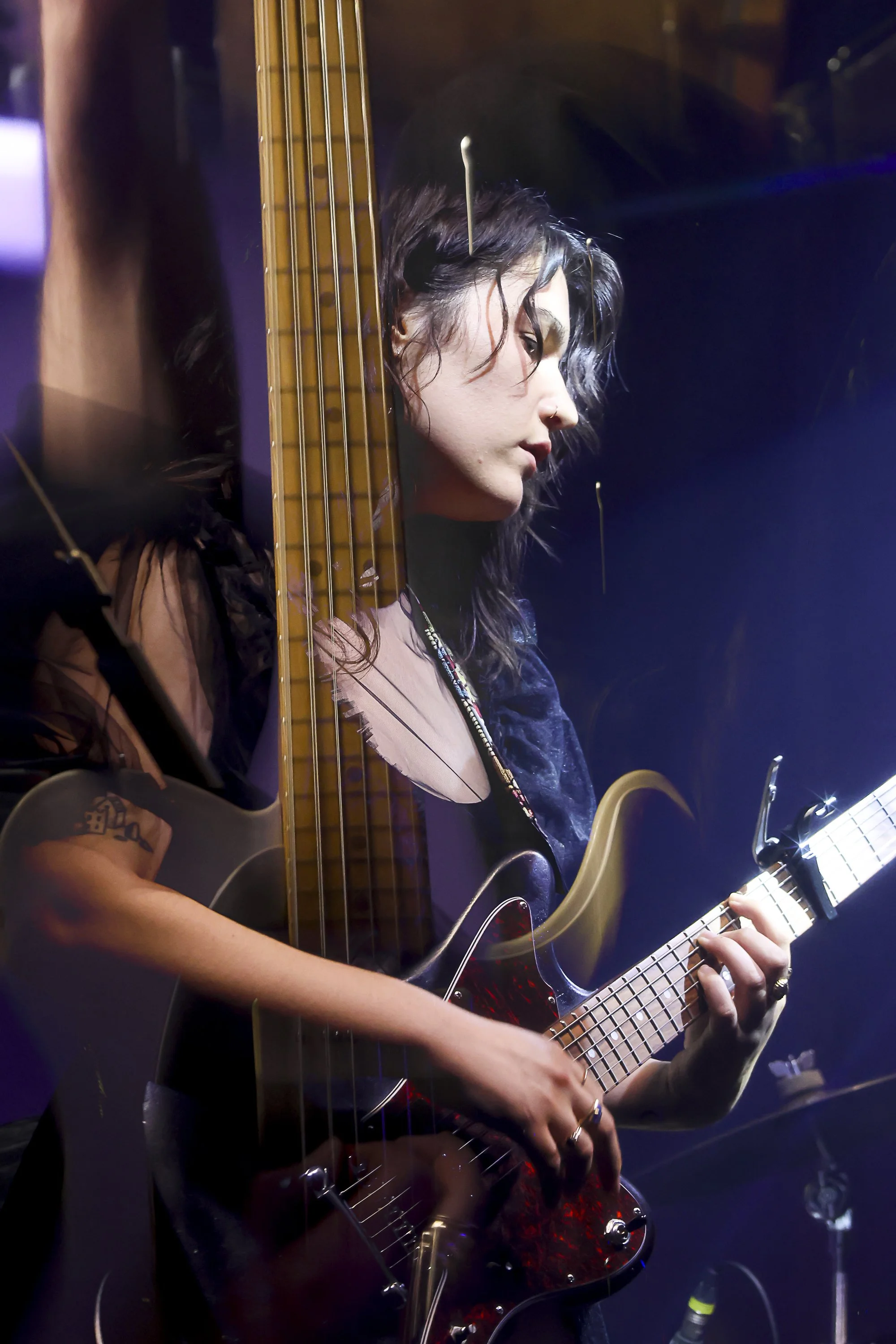 A woman with dark, curly hair partially covered by a guitar, playing an acoustic guitar on stage with blue and purple lighting.