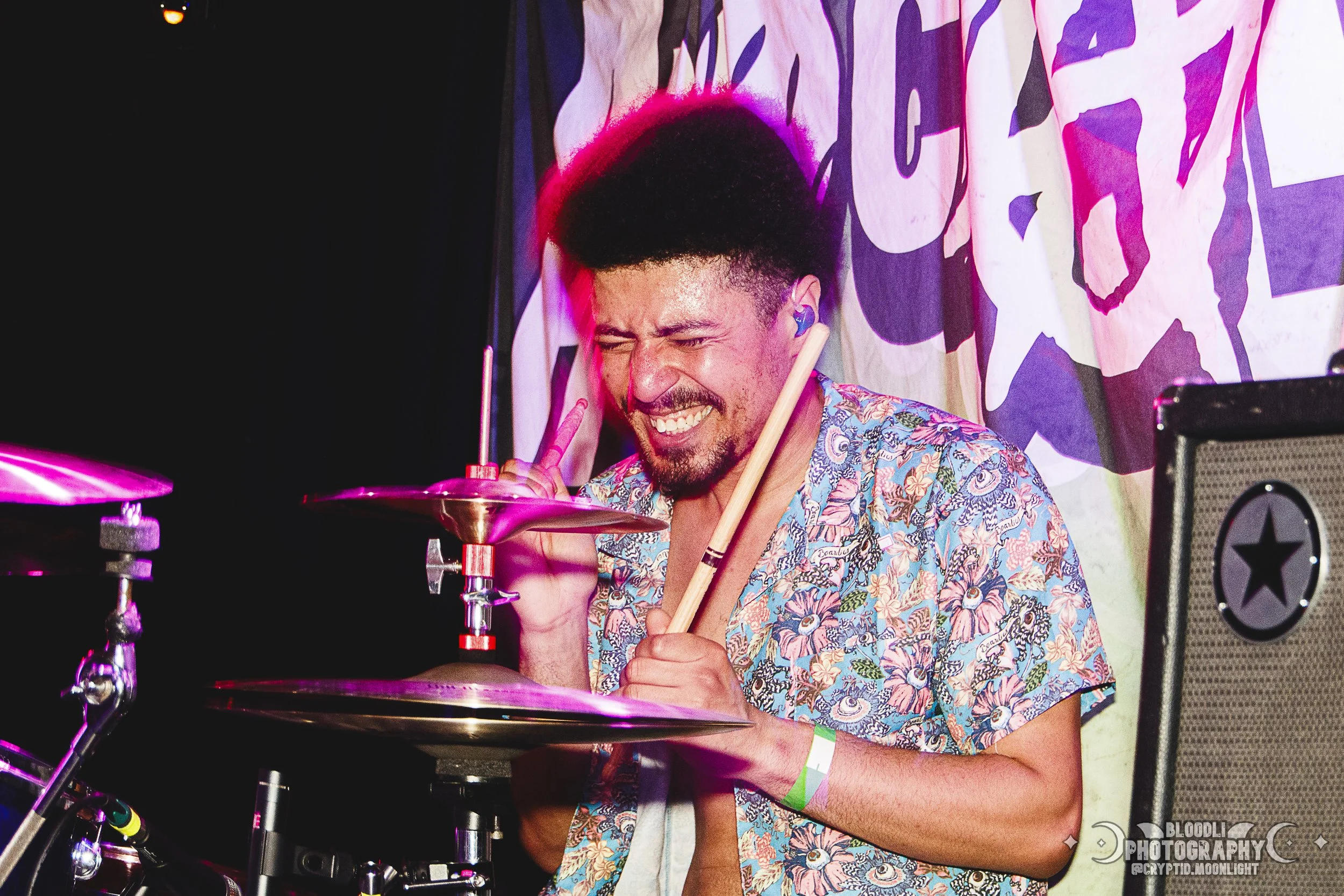 A young man with an afro hairstyle smiling and making a peace sign while playing drums during a live music performance.