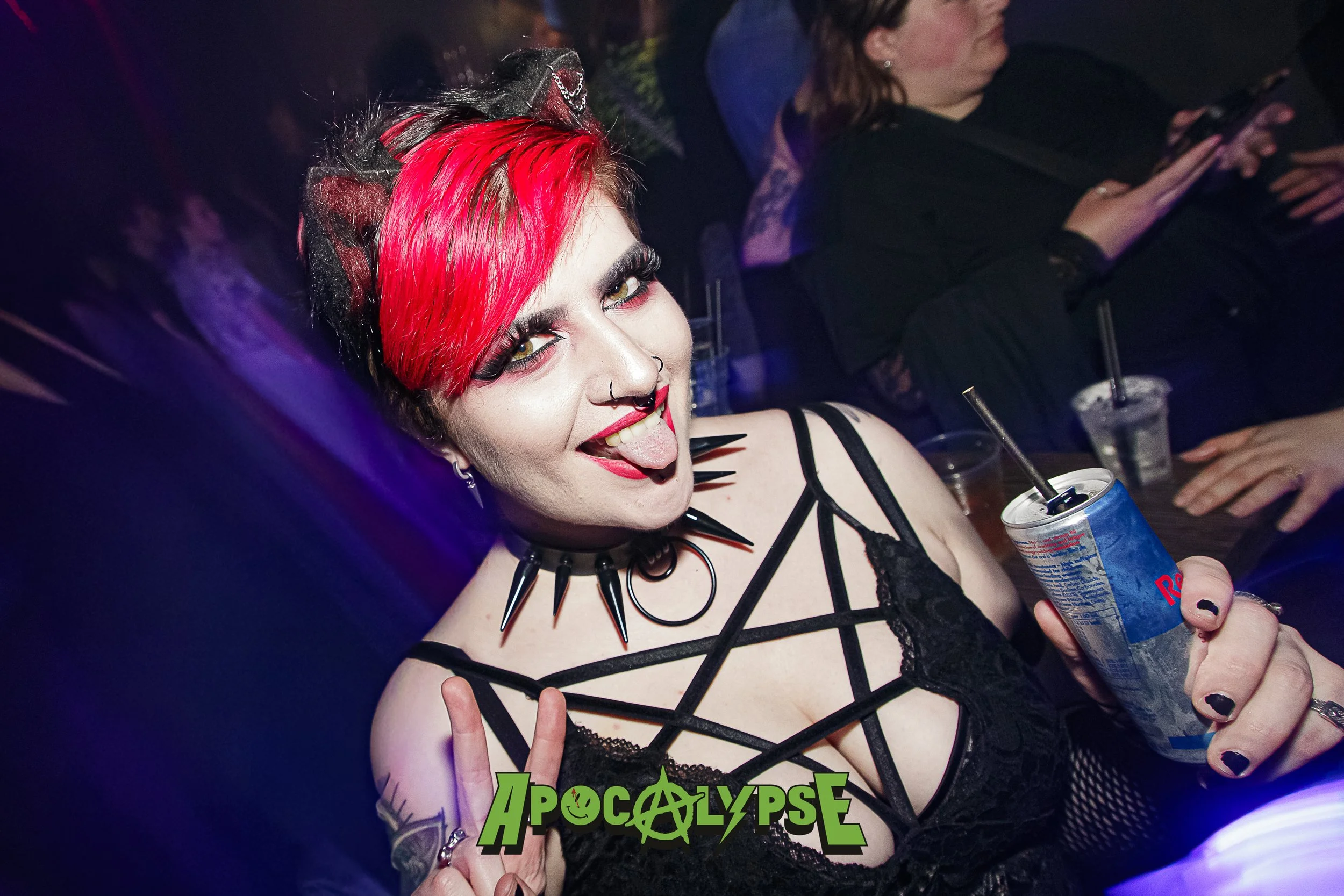 A woman with bright red hair, gothic makeup, and multiple piercings, wearing a black lace top and spiked jewelry, holding a can of soda with a straw in a dark, colorful party setting. She is making a peace sign and sticking out her tongue.