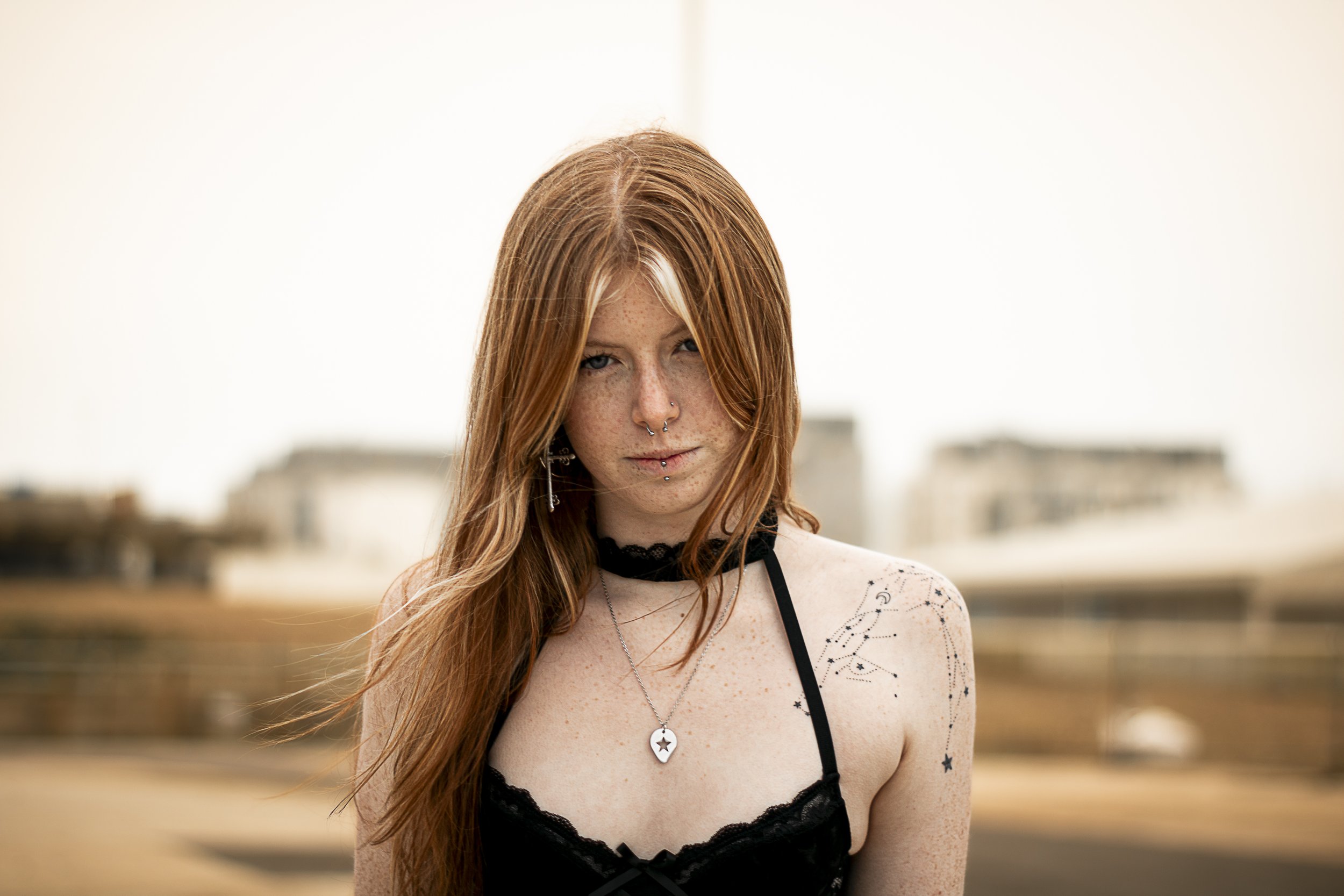 Red-haired woman with tattoos on shoulder and chest, wearing a black lace top, standing outdoors with blurred buildings in the background.