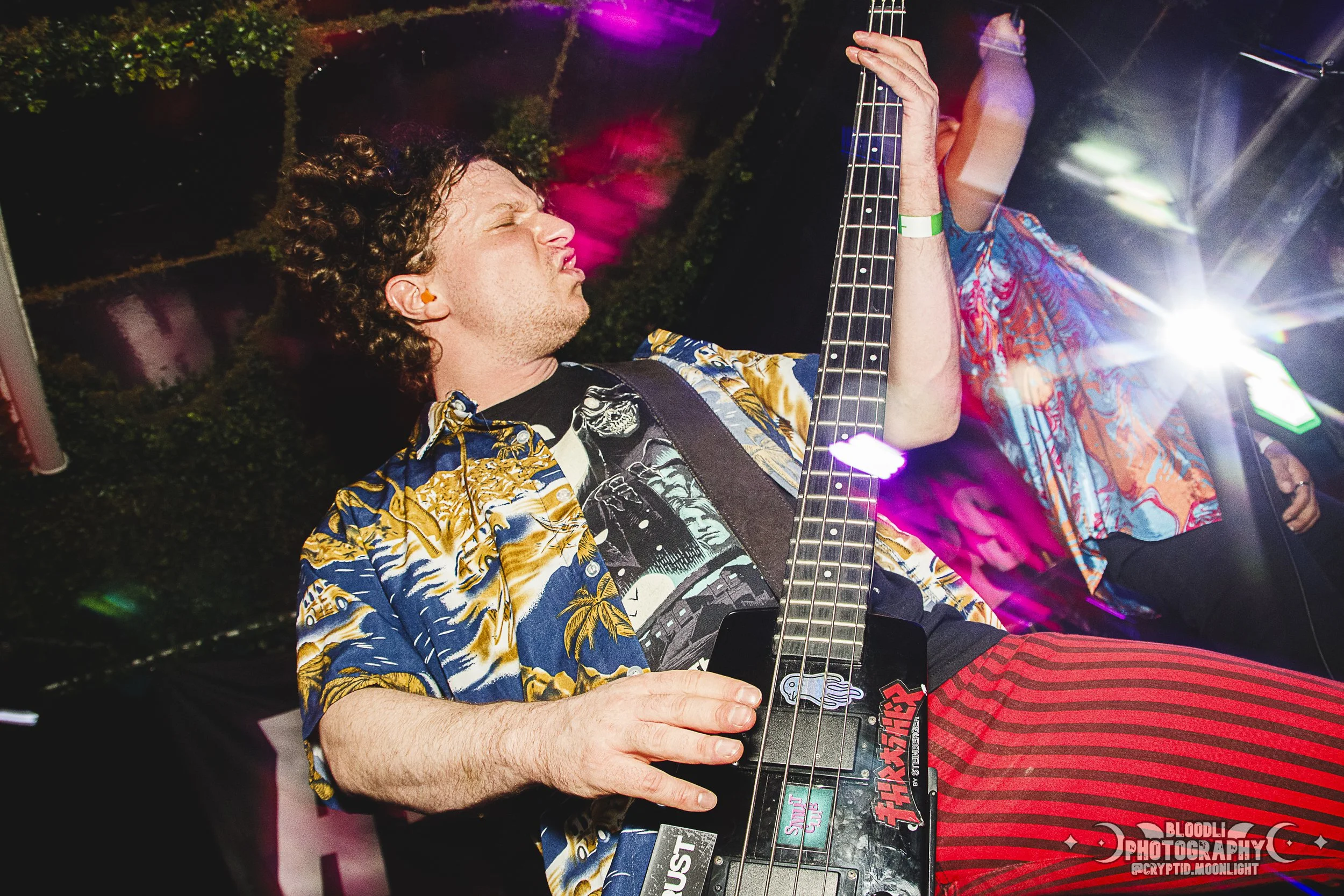 A man with curly hair playing an electric guitar on stage at a concert or event, wearing a colorful, patterned shirt and red striped pants, illuminated by stage lights.
